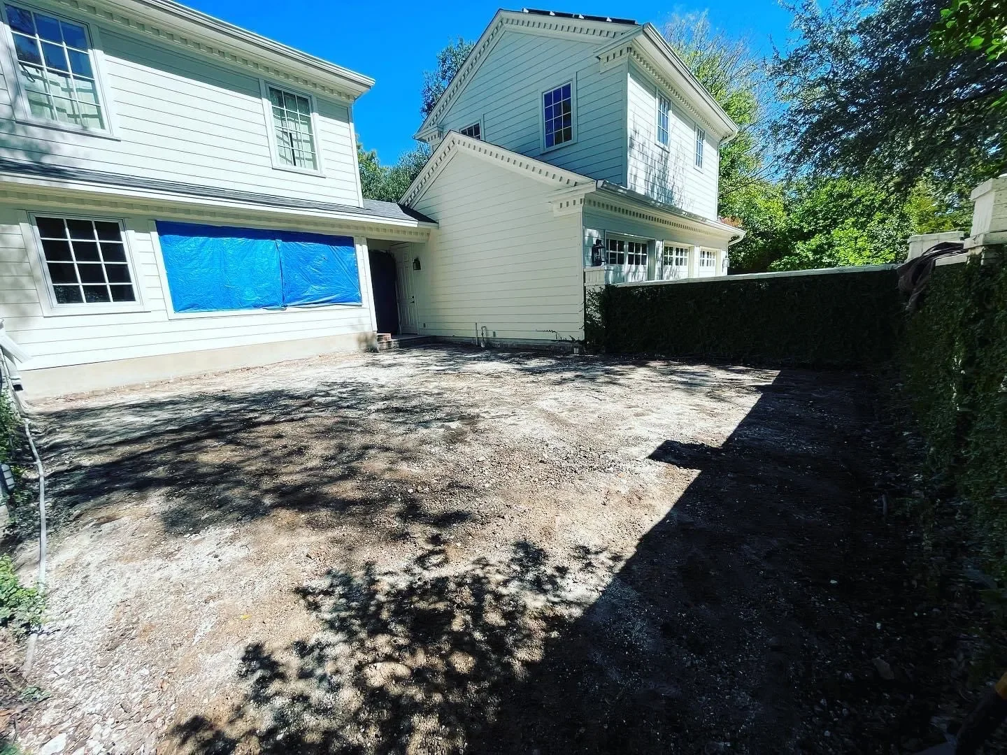 Backyard with dirt ground, shadowed areas, and two white houses with multiple windows, one house has a blue tarp covering a window, and the yard is surrounded by trees and greenery.