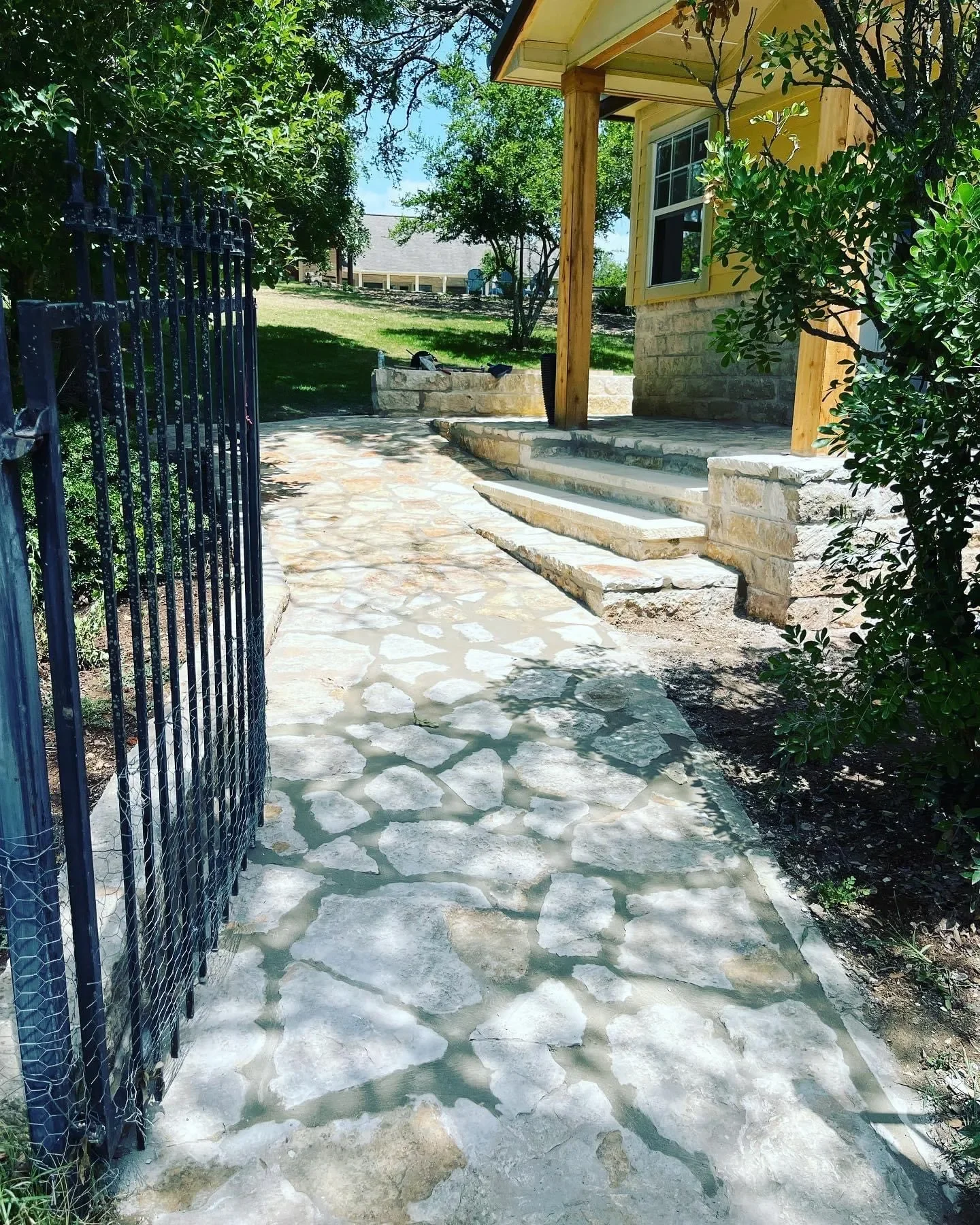 Stone pathway leading to a yellow house with a porch and steps, surrounded by bushes and trees, with a black gate on the left.