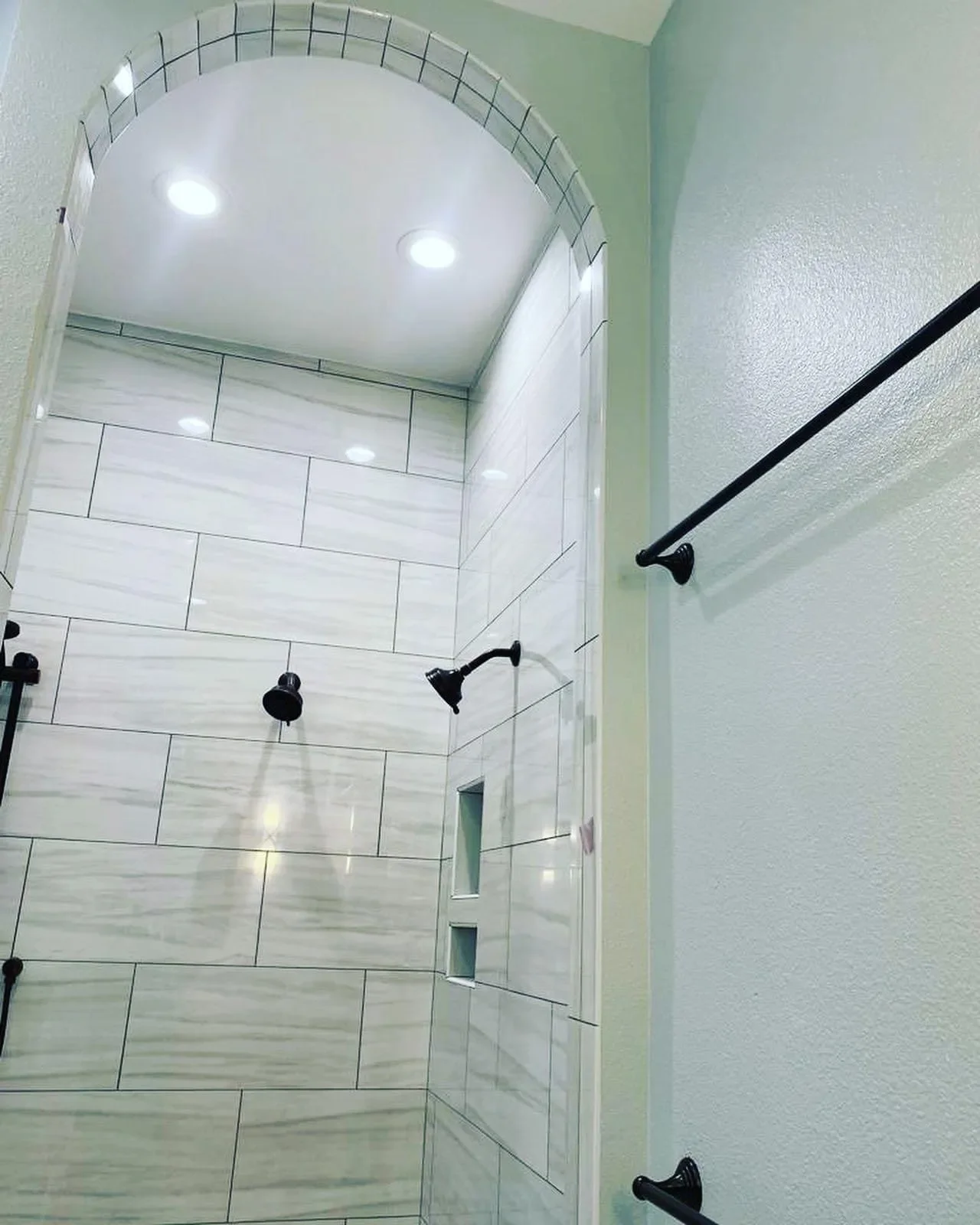 Interior of a modern shower with two black shower heads, white rectangular tiled walls, built-in wall niches, with a curved ceiling and recessed lighting.