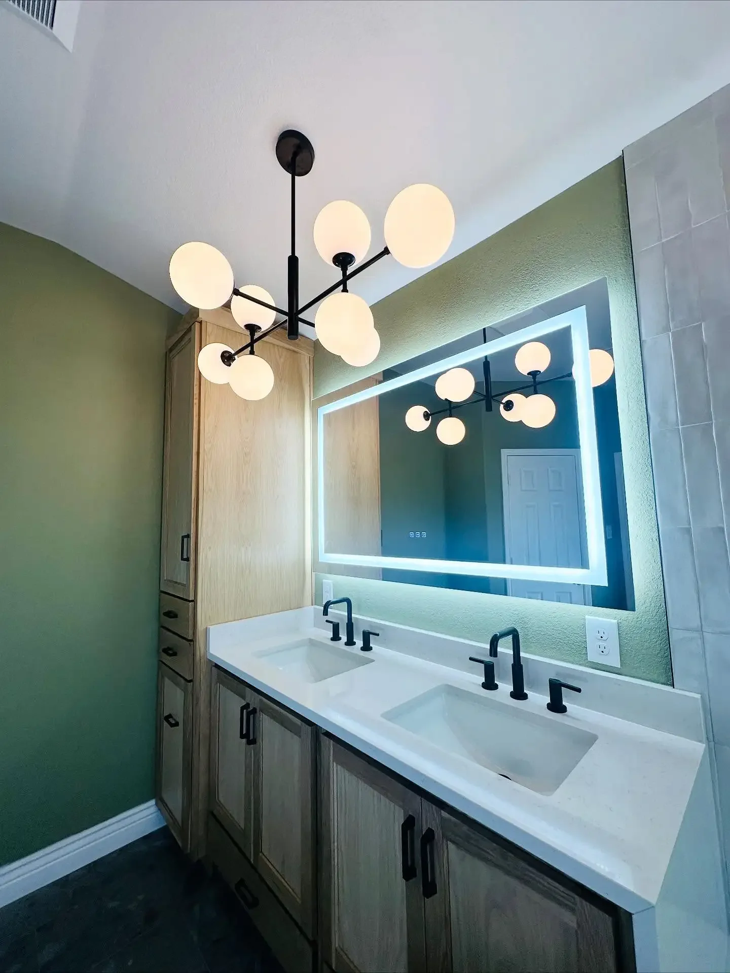 Modern bathroom with double sink vanity, illuminated mirror, black fixtures, wooden cabinet, green wall, and black ceiling chandelier with globe light bulbs.