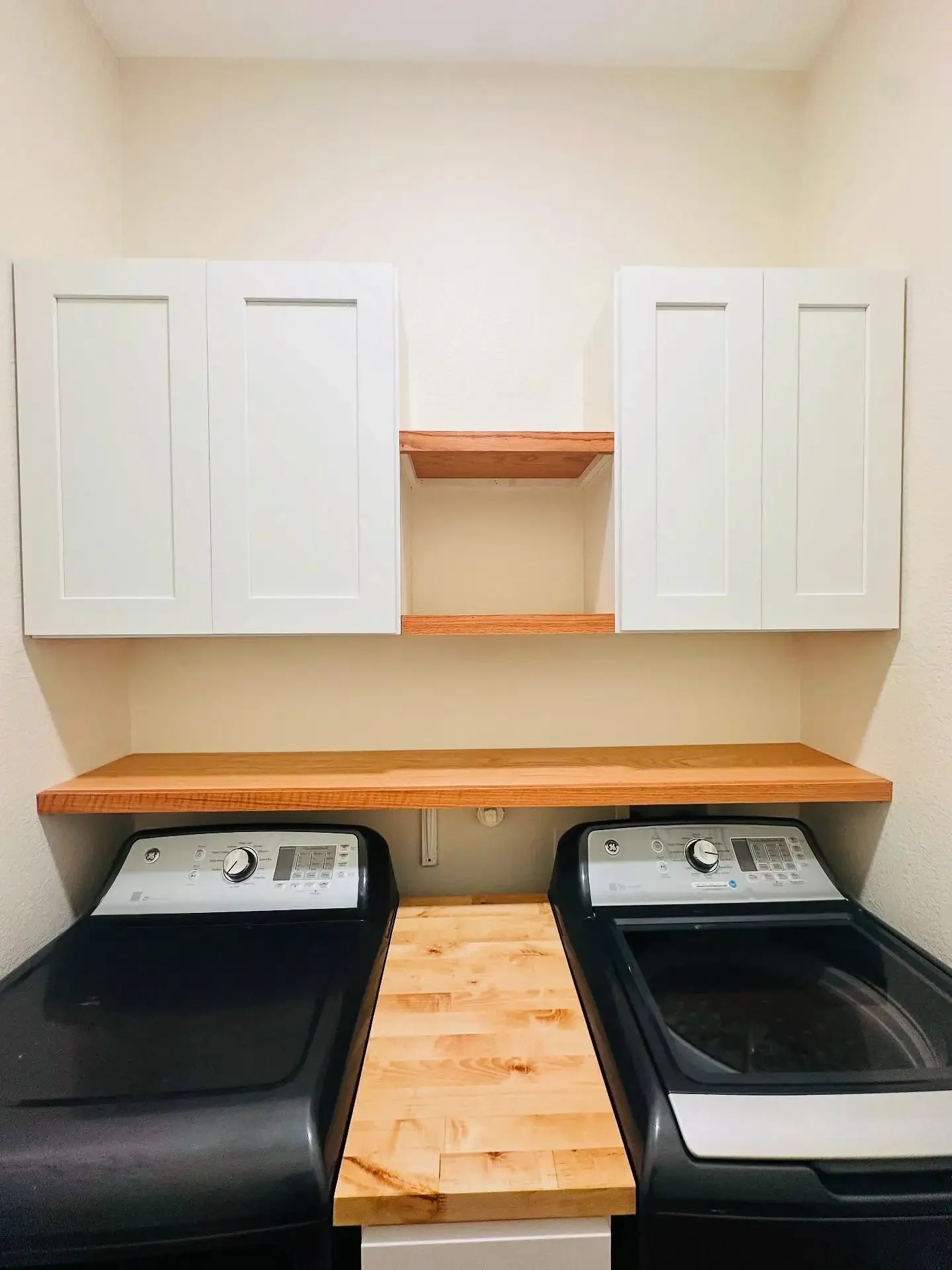 A laundry room with two black washing machines, white cabinets above, and a wooden countertop between them.