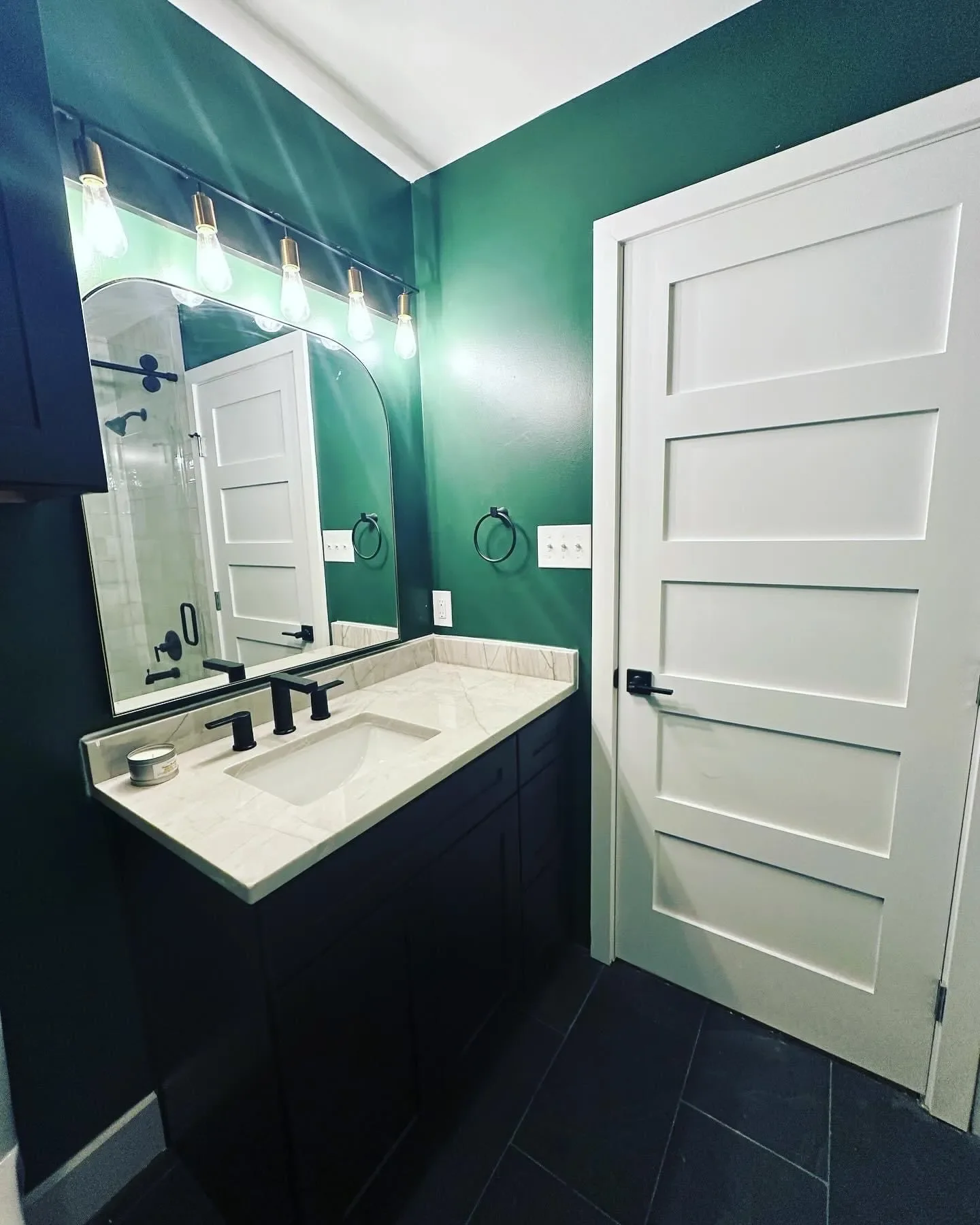 Modern bathroom with dark green walls, white door with horizontal paneling, black vanity with marble countertop, large mirror, and black fixtures, including black faucet and towel rings.