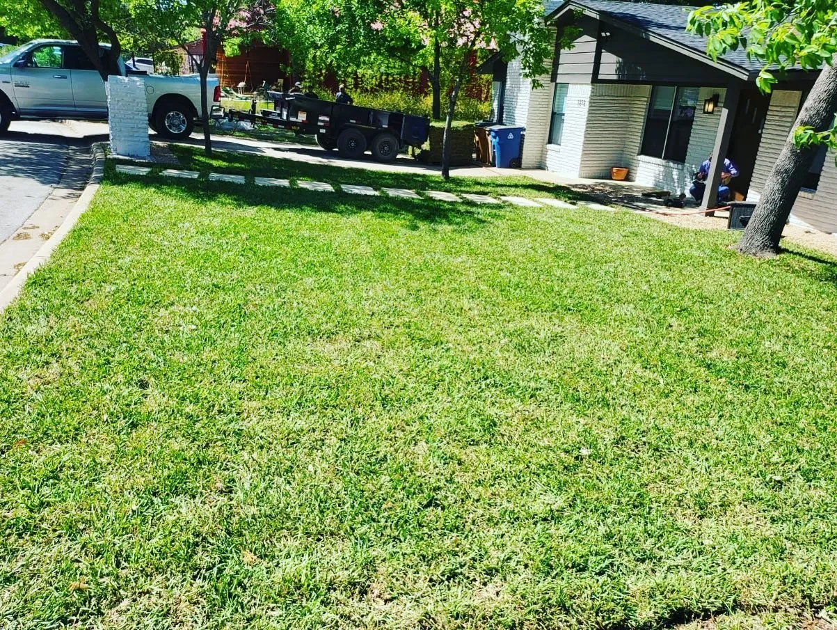 A front yard with green grass, a sidewalk, trees, a house with a porch, parked vehicles, and a trailer in the driveway on a sunny day.
