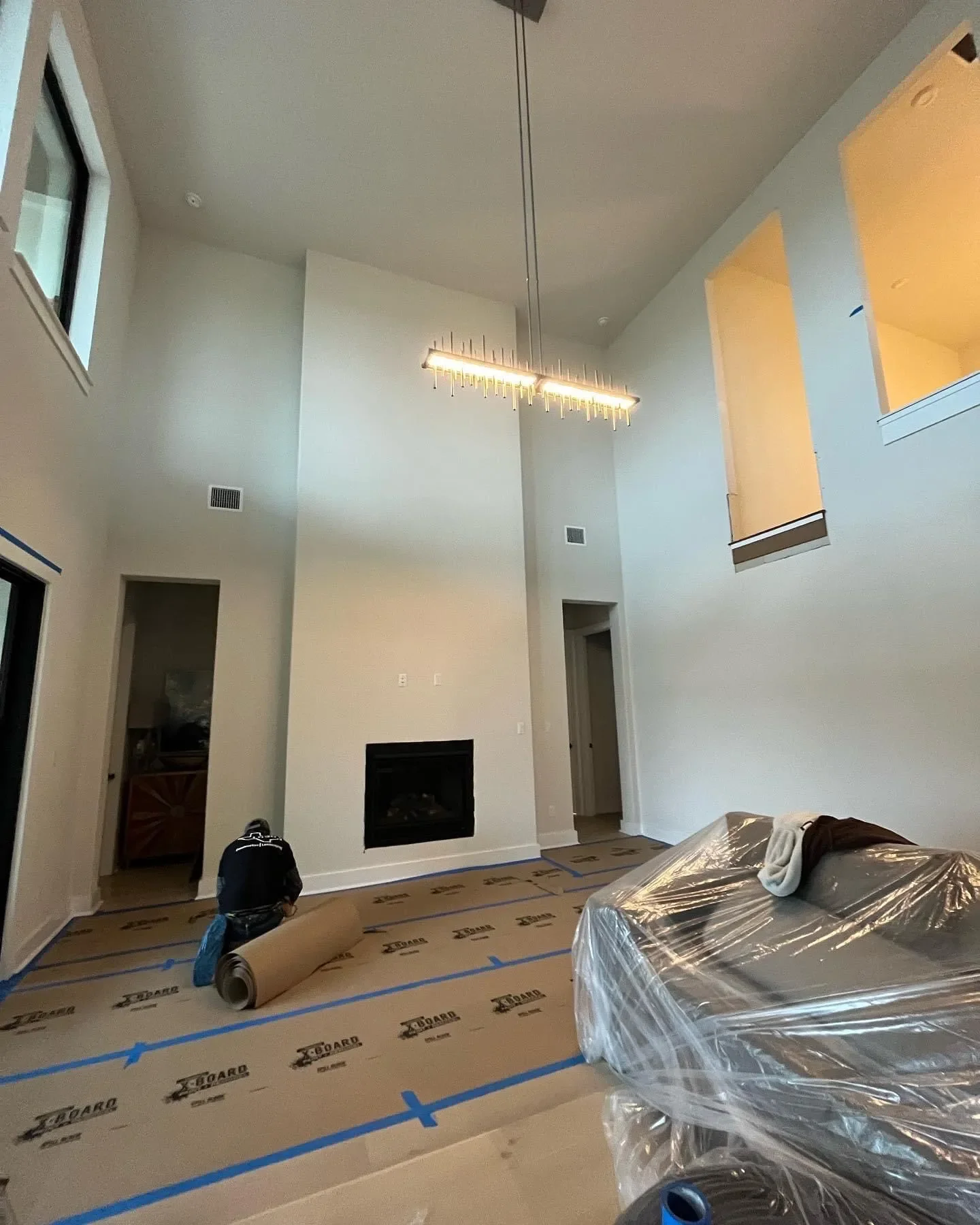 Living room under construction with a worker laying down carpet, large windows, a modern chandelier, and a white fireplace.