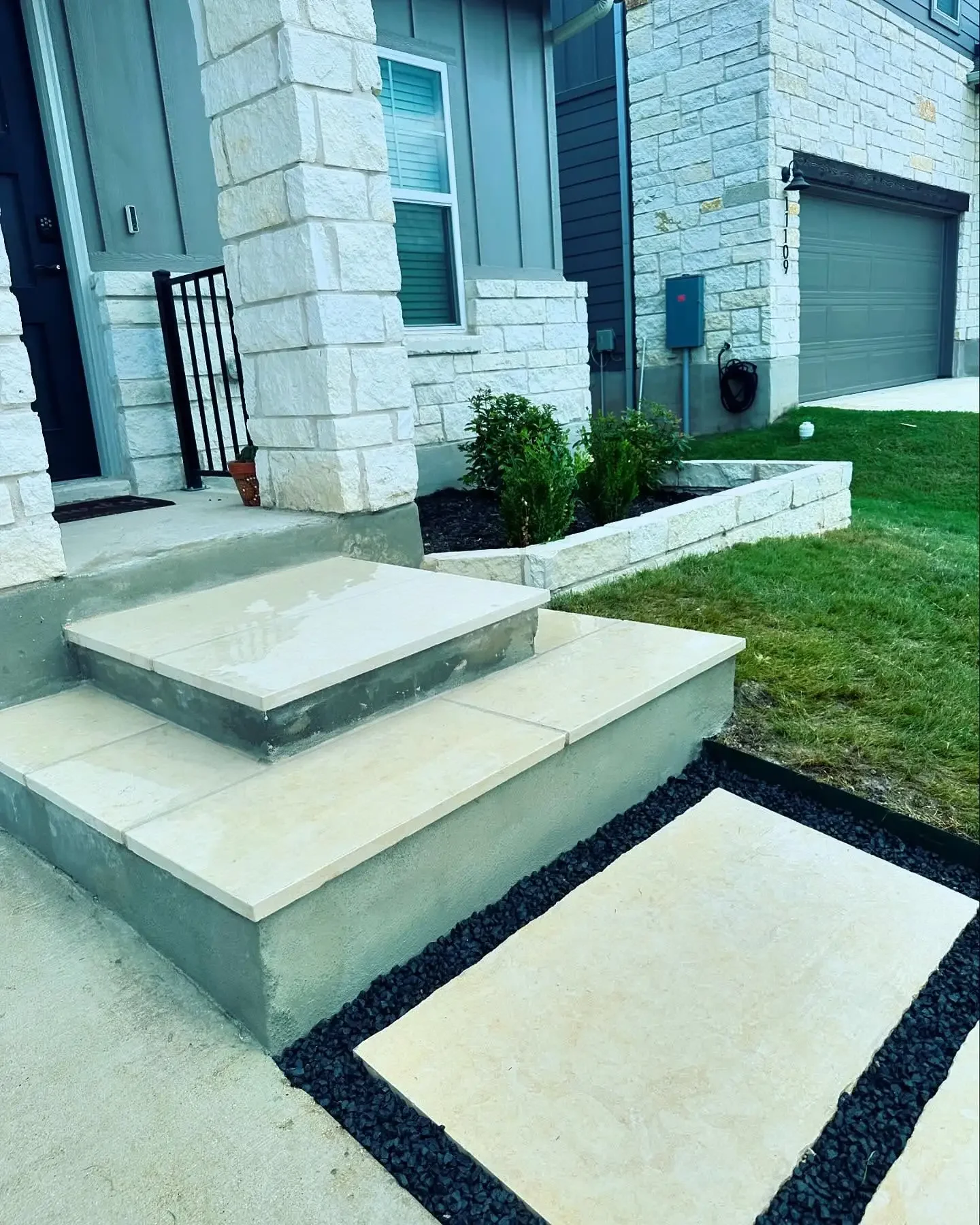 Front entrance of a house with white stone steps, a small garden bed with green plants, a stone walkway bordered with black gravel, a green garage door, and a lawn.