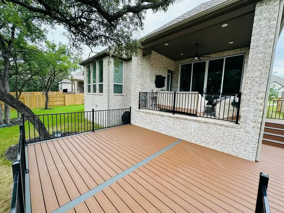 Backyard with a wooden deck, black metal railing, and a covered porch with outdoor furniture inside a brick house with large windows.