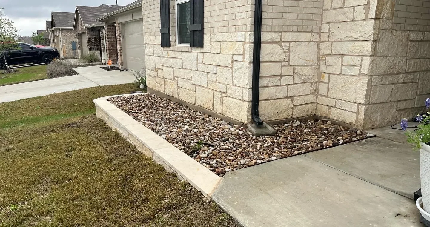 Front yard of a house with a small bed of rocks bordered by concrete, next to a light-colored brick wall with a black downspout and window with black shutters. The sidewalk is concrete, and there is a grass lawn nearby.