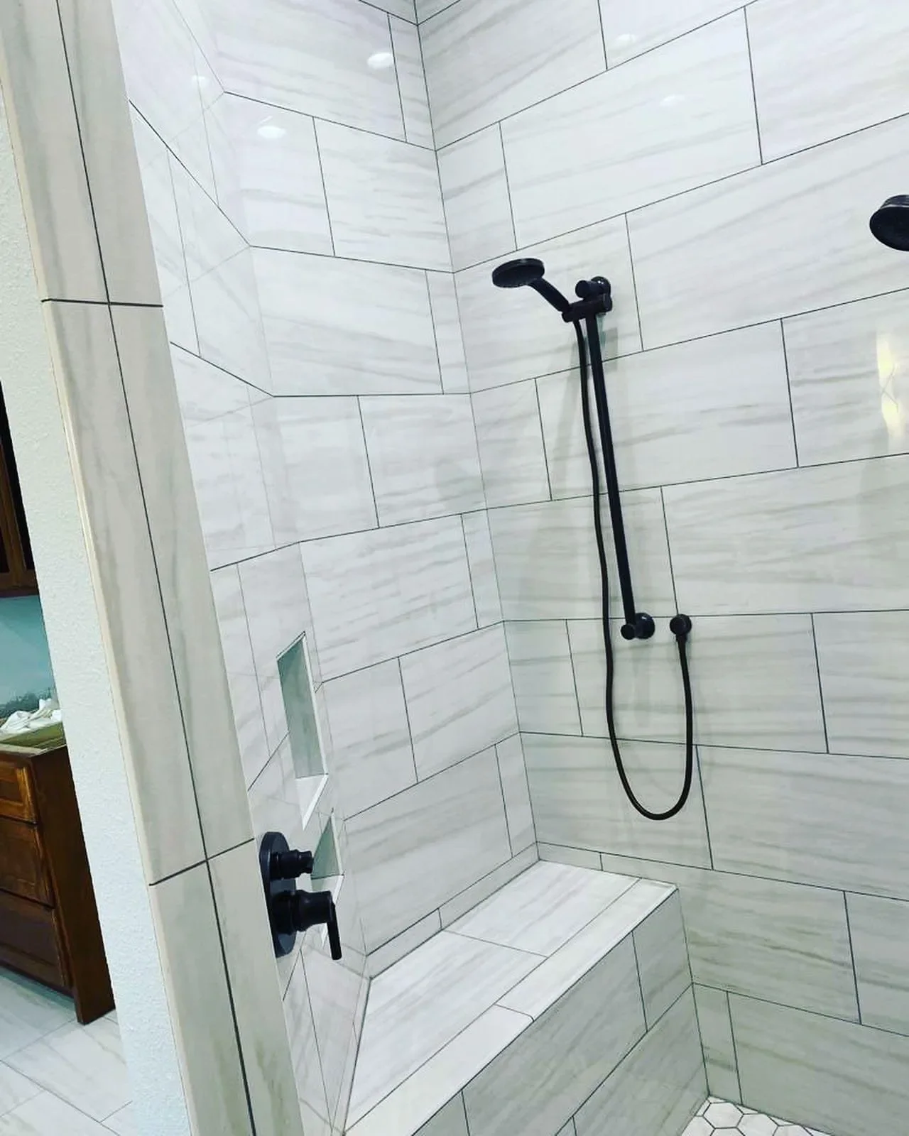 Bathroom shower with white tiled walls, black handheld showerhead, and built-in bench.