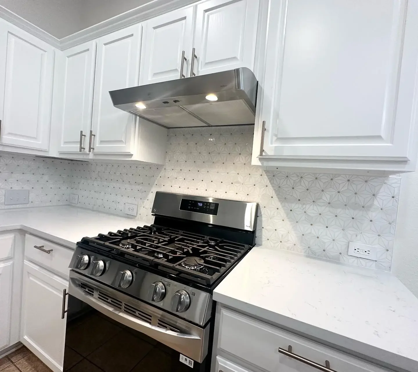 Kitchen with white cabinets, white countertops, and a stainless steel gas stove with control knobs. There is a stainless steel range hood above the stove with built-in lights. The backsplash features white tiles with geometric patterns. Multiple elec