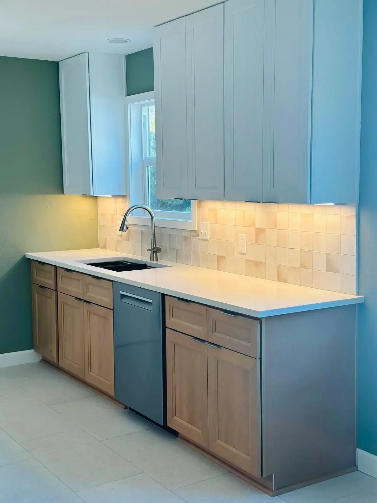 Modern kitchen with beige lower cabinets, light blue upper cabinets, a white countertop, and a window above the sink.