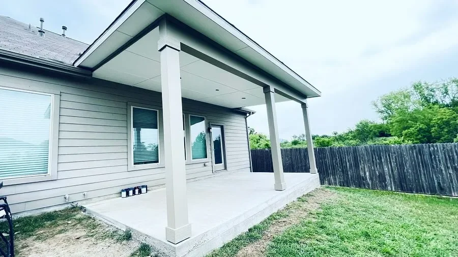 Backyard patio of a house with a covered porch supported by four white columns, a concrete floor, and a wooden fence in the background. The house has light gray siding and two large windows.
