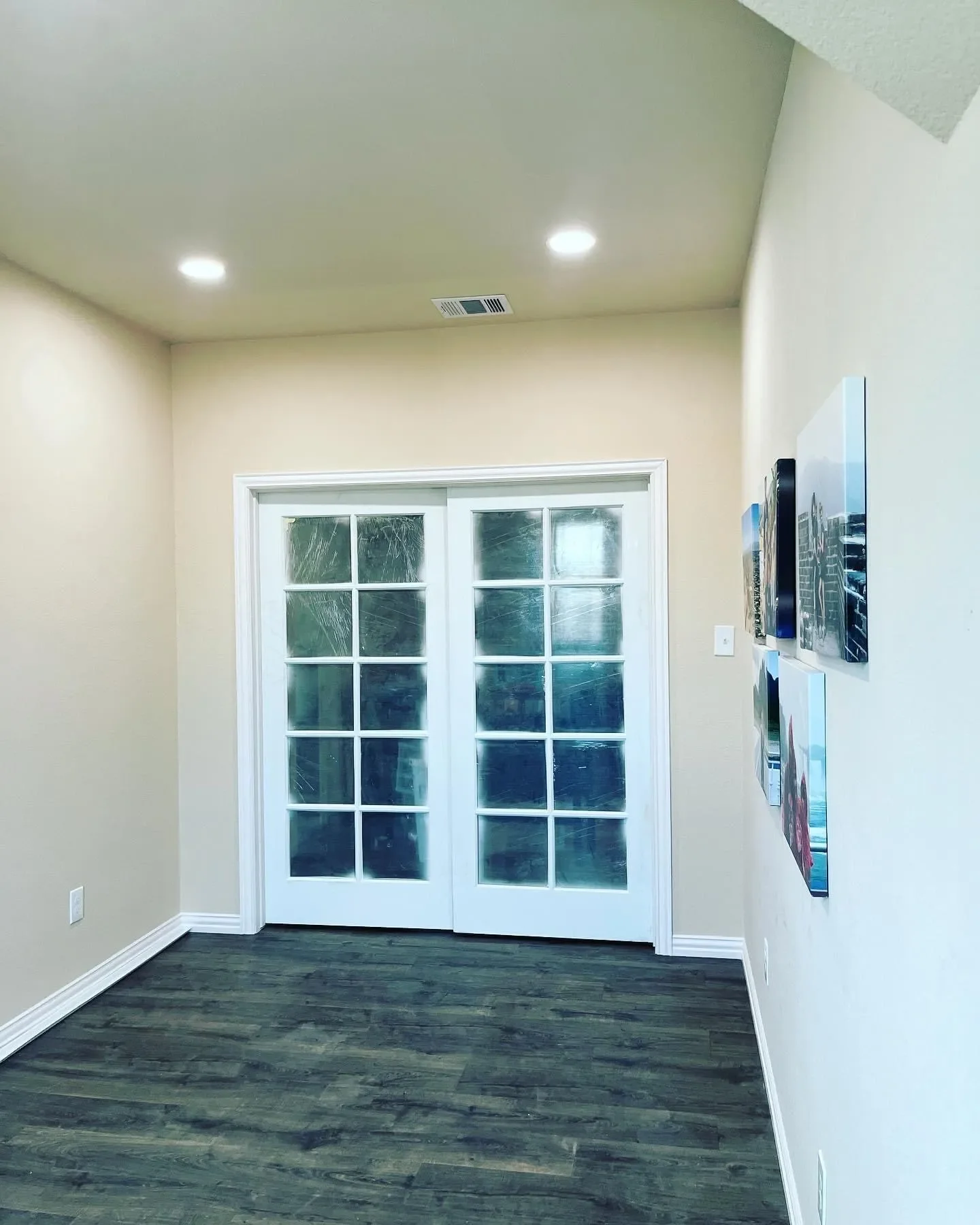 A room with hardwood floors, beige walls, a pair of white glass-paneled double doors, and a wall-mounted TV with pictures on the right wall. There are two ceiling lights and a vent in the ceiling.