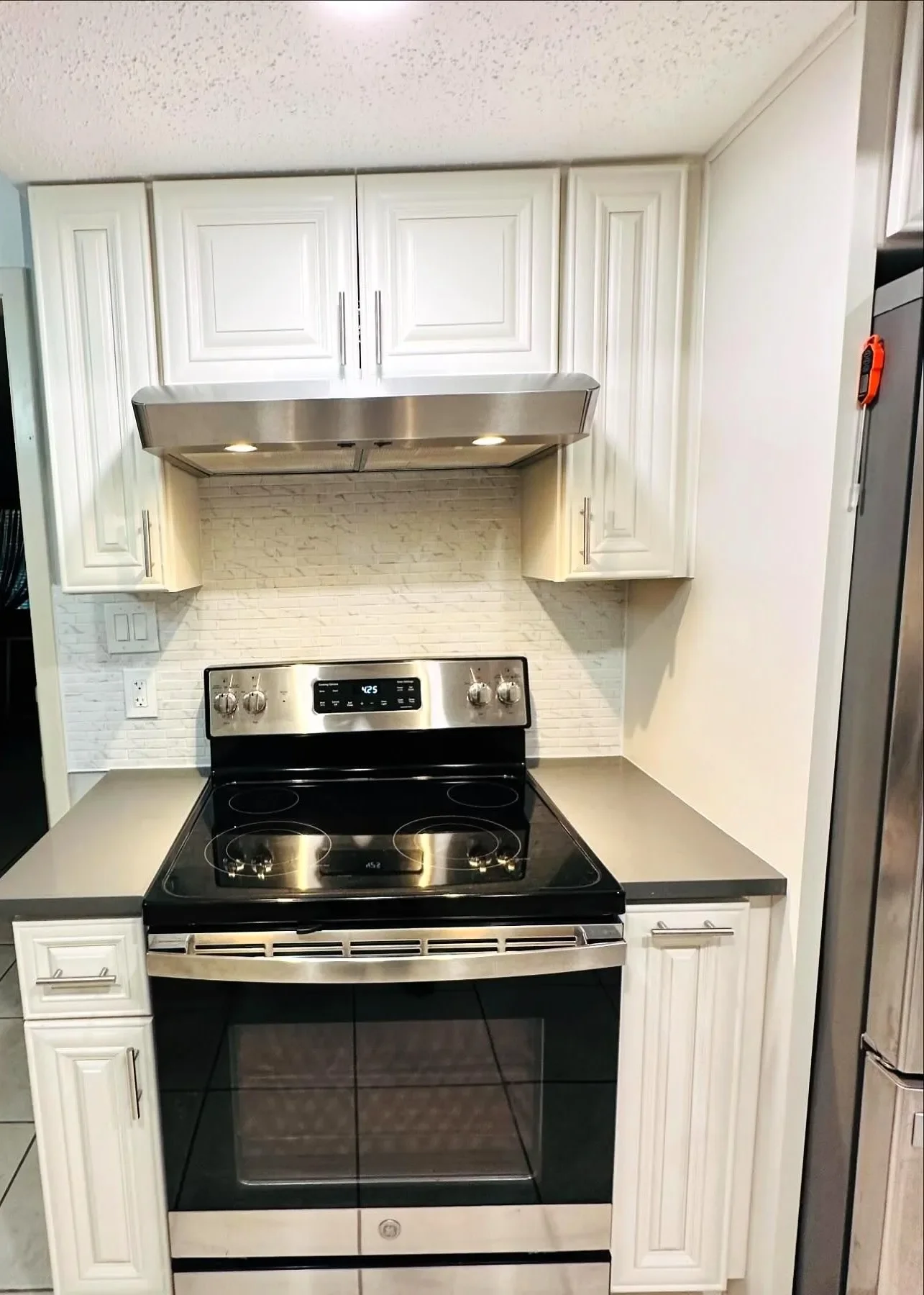 Kitchen with white cabinets, stainless steel stove and range hood, black glass cooktop, and grey countertop. Small section of tile backsplash behind the stove.