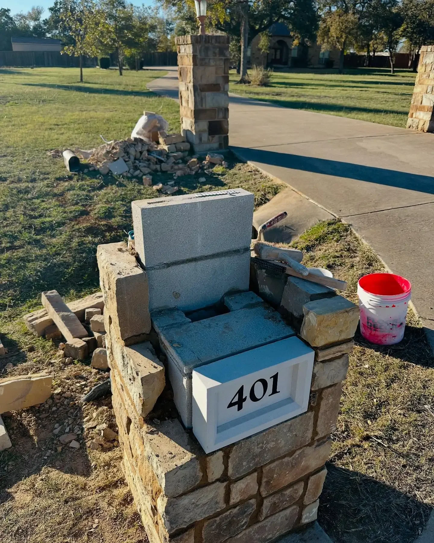 Close-up of a brick mailbox at 401, with construction materials and tools around it, including a bucket, a trowel, and a small pile of bricks. The background shows a sidewalk, a stone pillar, trees, and a house.