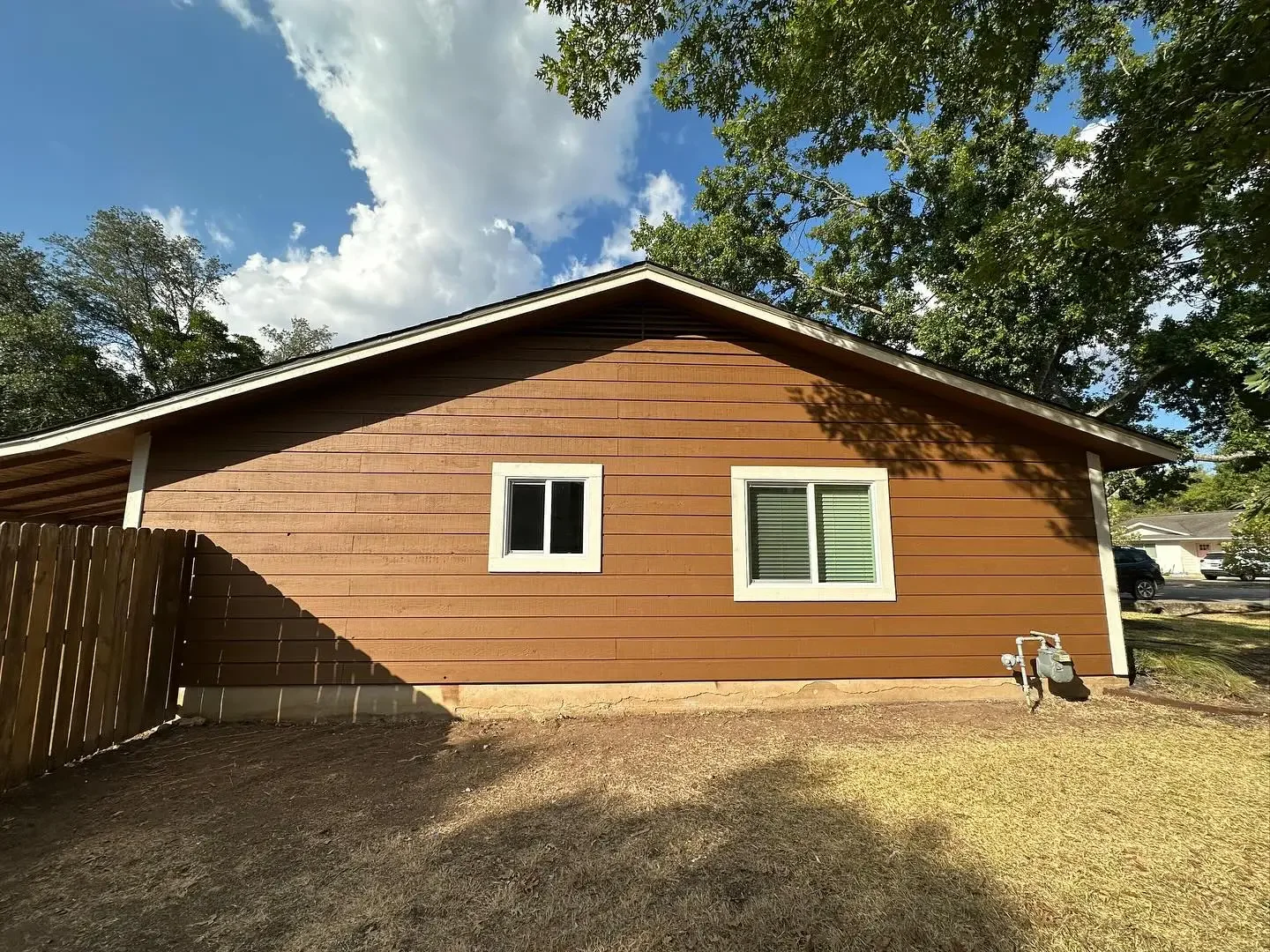 Back of a brown house with two white-framed windows, a wooden fence, and a small grassy area. There are trees overhead and a partly cloudy sky.