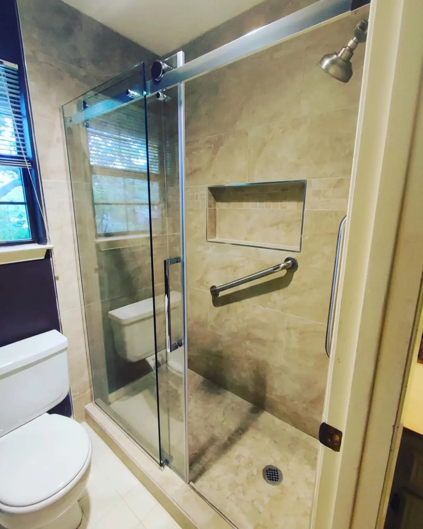 Bathroom shower with glass enclosure, beige tiled walls, built-in niche, grab bar, and a showerhead. Part of a toilet and window are visible.