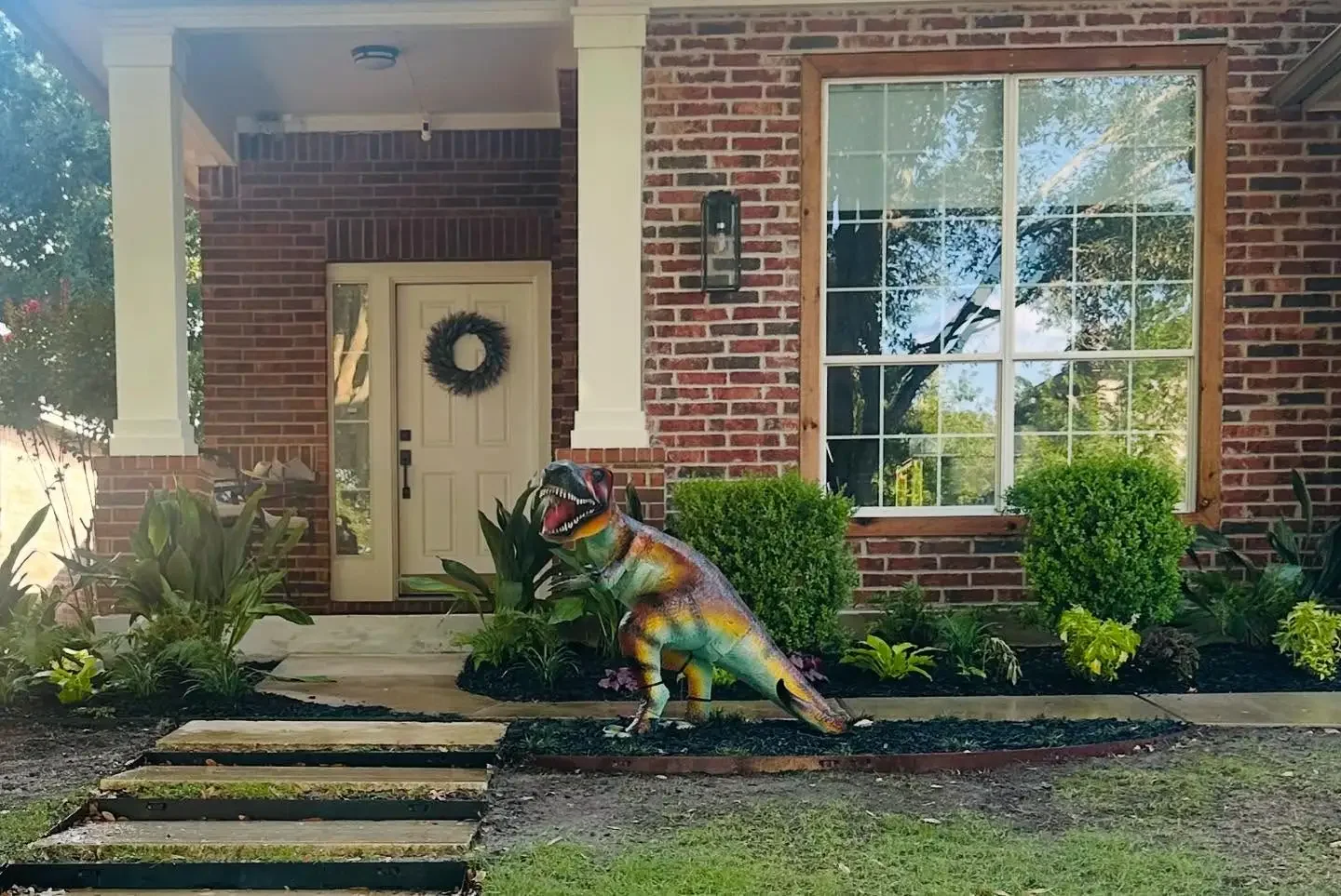A decorated front yard of a brick house with a dinosaur statue in the garden.