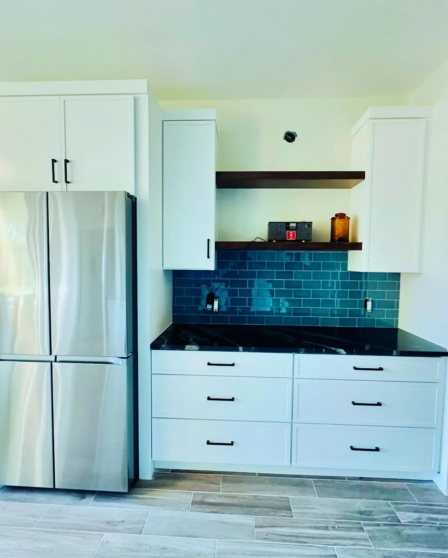 Modern kitchen with a stainless steel refrigerator, white cabinets, black countertop, blue tile backsplash, and wooden shelves.