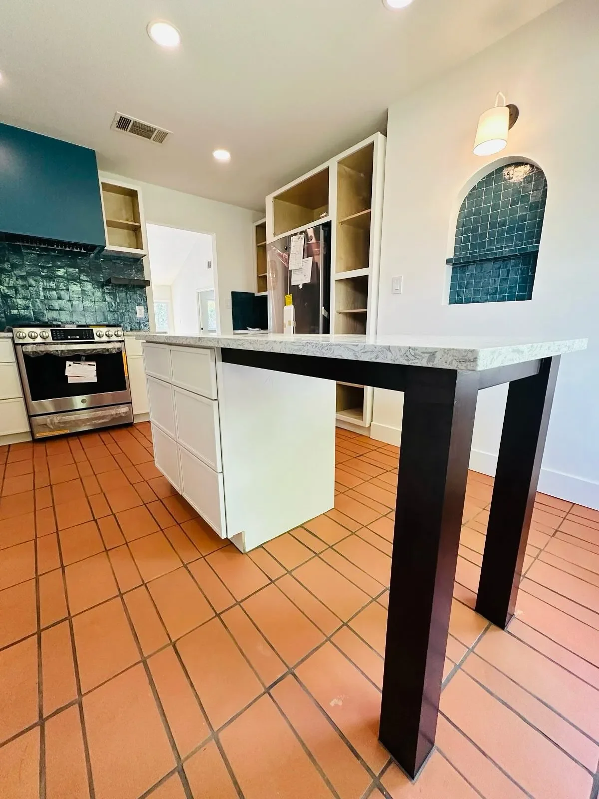 Inside a kitchen showing a white island with a marble countertop, open shelves, a black and stainless steel oven, and a blue tiled wall with a small arched niche.