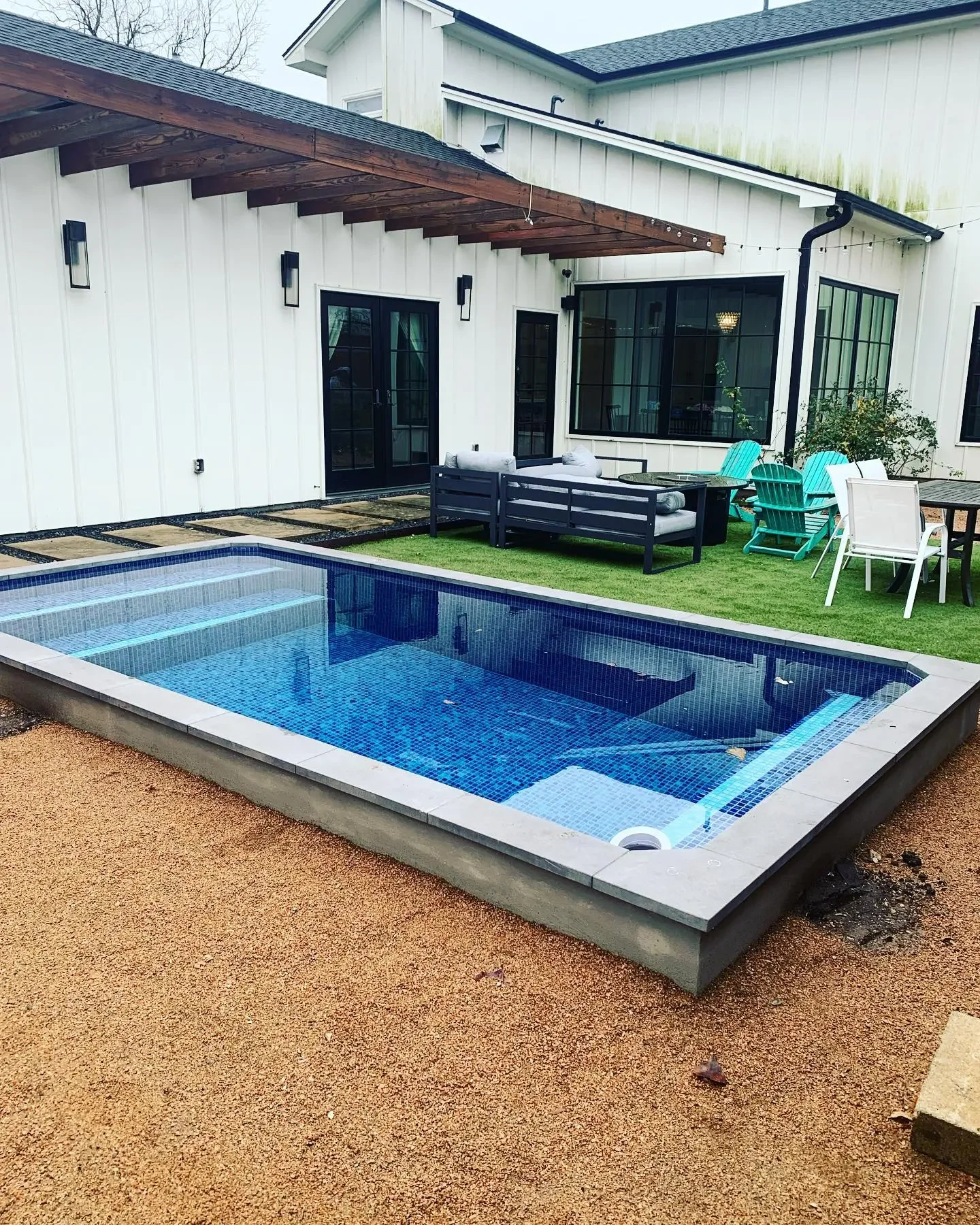 Backyard with small swimming pool, outdoor furniture including a gray sofa, teal and white chairs, and a glass table, patio stones, and a white house with black doors and large windows.