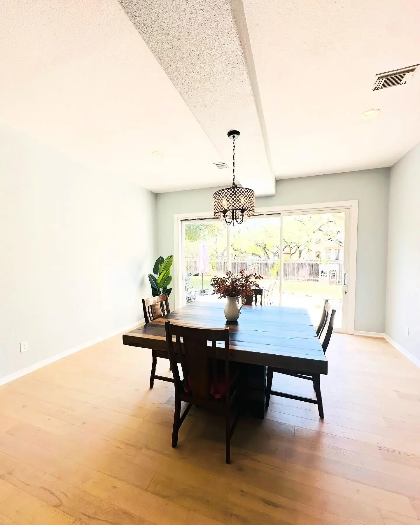 Dining room with a wooden table, six chairs, a chandelier, a sliding glass door, a potted plant, and a vase with flowers.