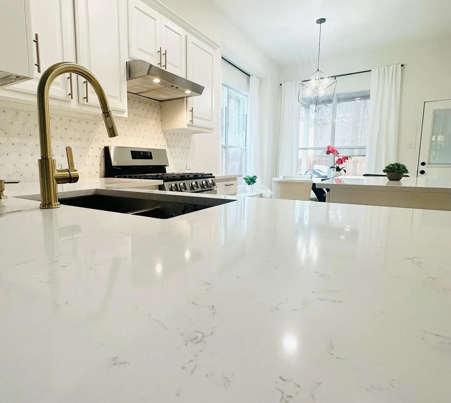 Modern kitchen with white cabinets, a gold faucet, a black sink, stainless steel stove, and natural light from large windows with white curtains.