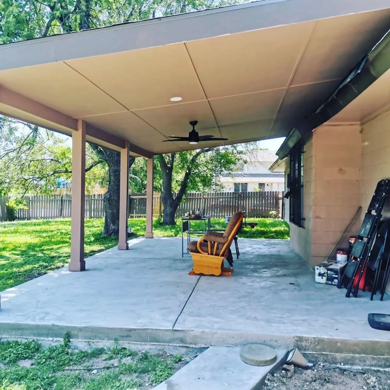 Covered patio with a ceiling fan, a rocking chair, a small table, and gardening supplies, with trees and a wooden fence in the background.