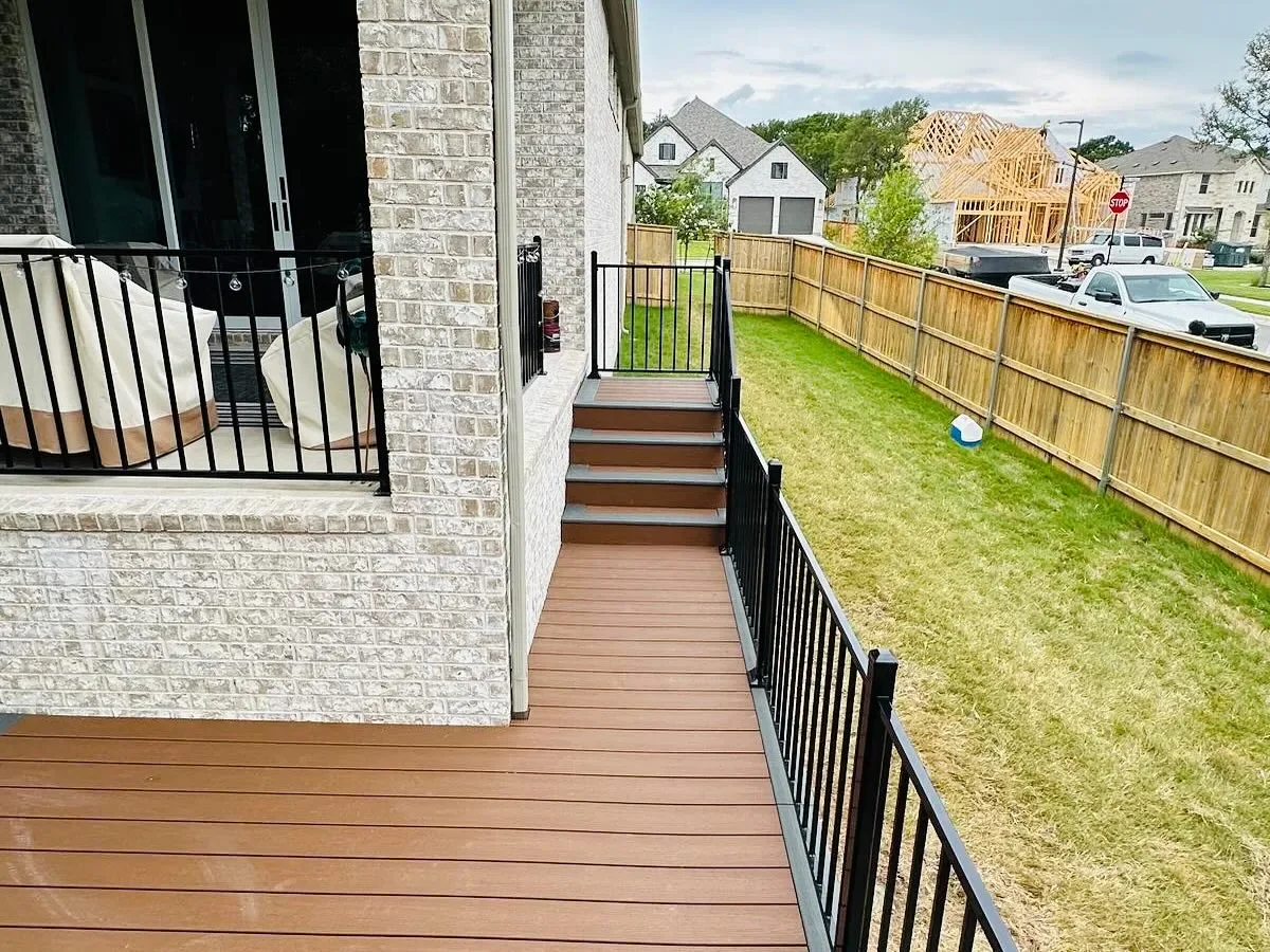 A backyard patio with wooden flooring and black metal railing, outdoor steps leading to the grass yard, and a brick house wall with sliding glass doors, neighboring houses, some under construction, and a wooden fence.