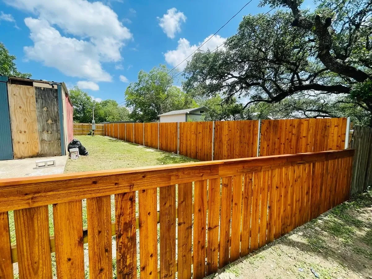 View of a backyard with a wooden fence, a grassy area, and a shed. There are trees and a partly cloudy sky.
