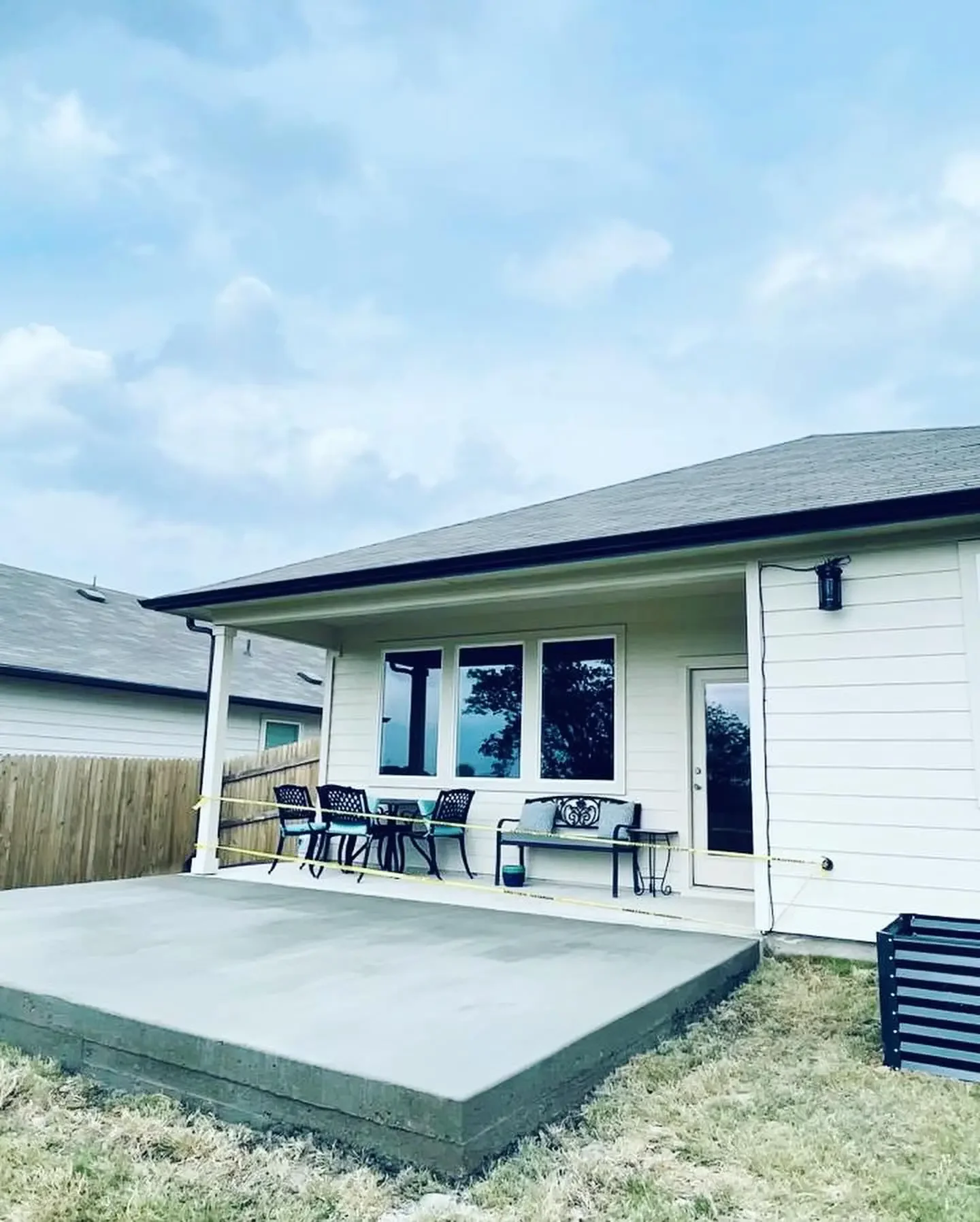 Backyard patio with outdoor seating, a bench, and a yellow caution tape around it, next to a white house with two large windows and a door, fenced backyard, overcast sky.