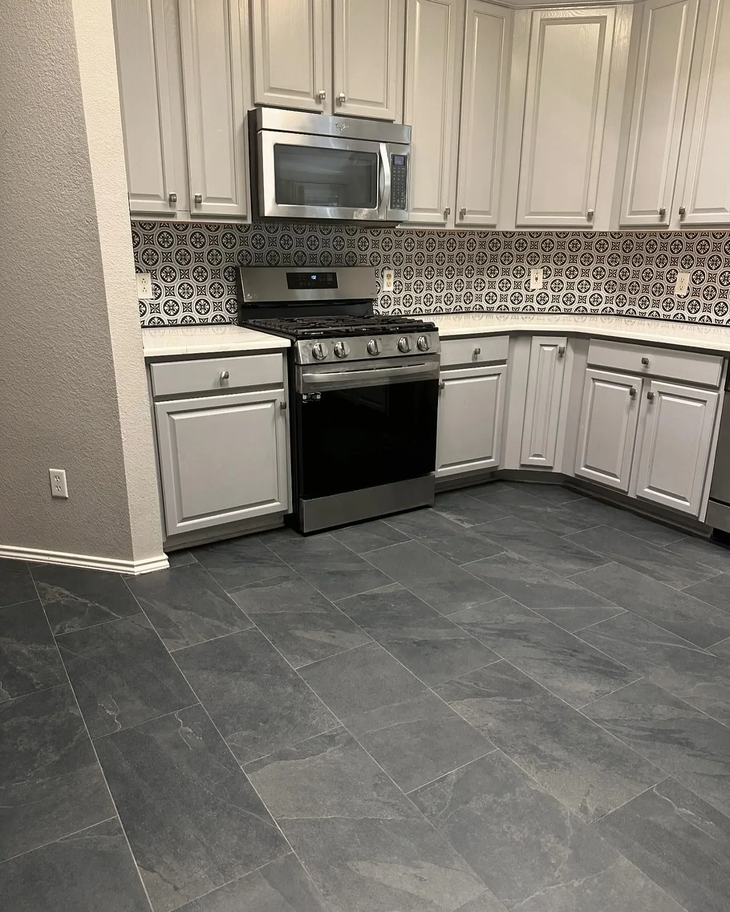 Kitchen with gray cabinets, stainless steel microwave and stove, black and white patterned backsplash, gray tile floor.