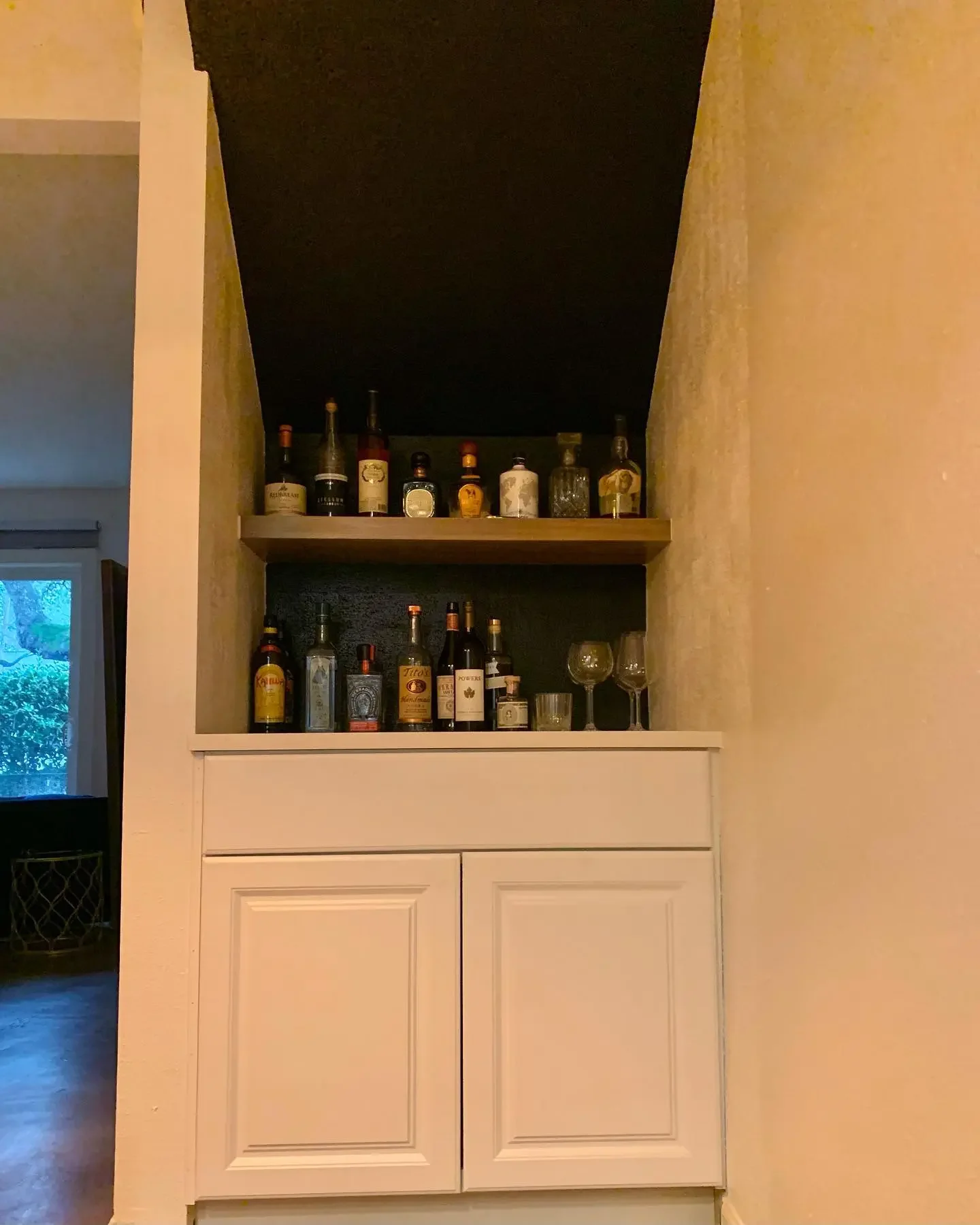 A built-in bar area with a white cabinet below and two open shelves above, holding various bottles of liquor and some glasses, located in a home interior.