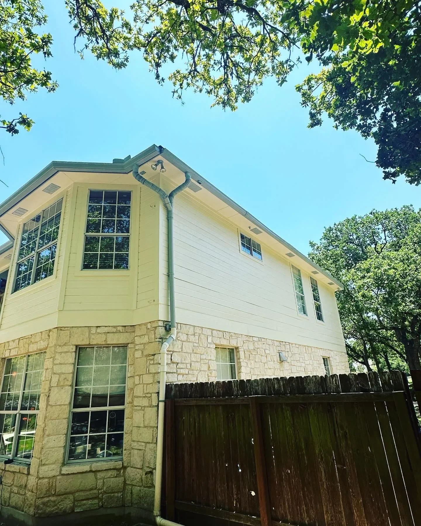 A two-story house with a stone foundation and white siding, surrounded by trees, with a wooden fence in the foreground and a bright blue sky overhead.