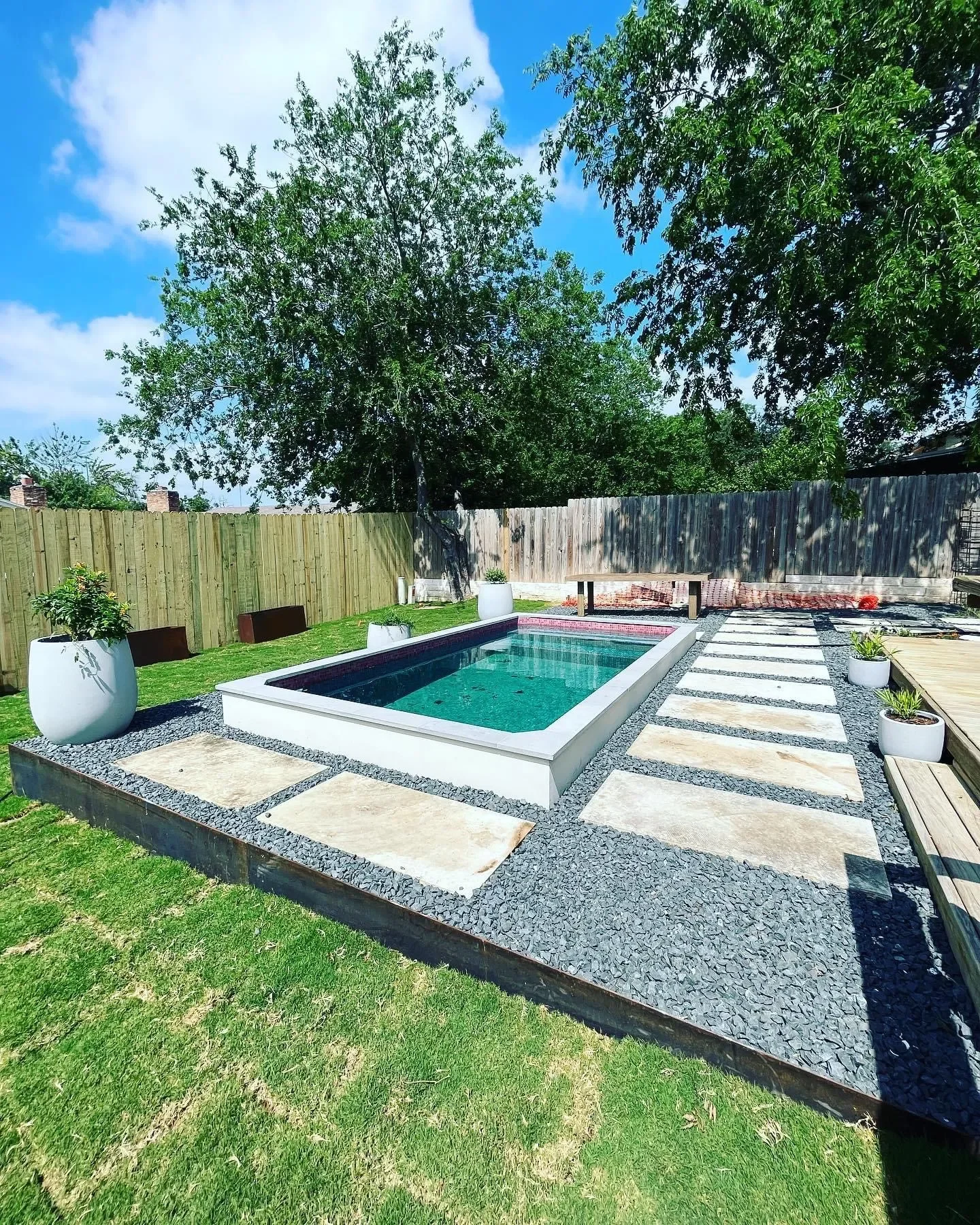 Backyard with a rectangular swimming pool, potted plants, stepping stones, a wooden deck, grassy area, tall trees, and a wooden fence under a partly cloudy sky.