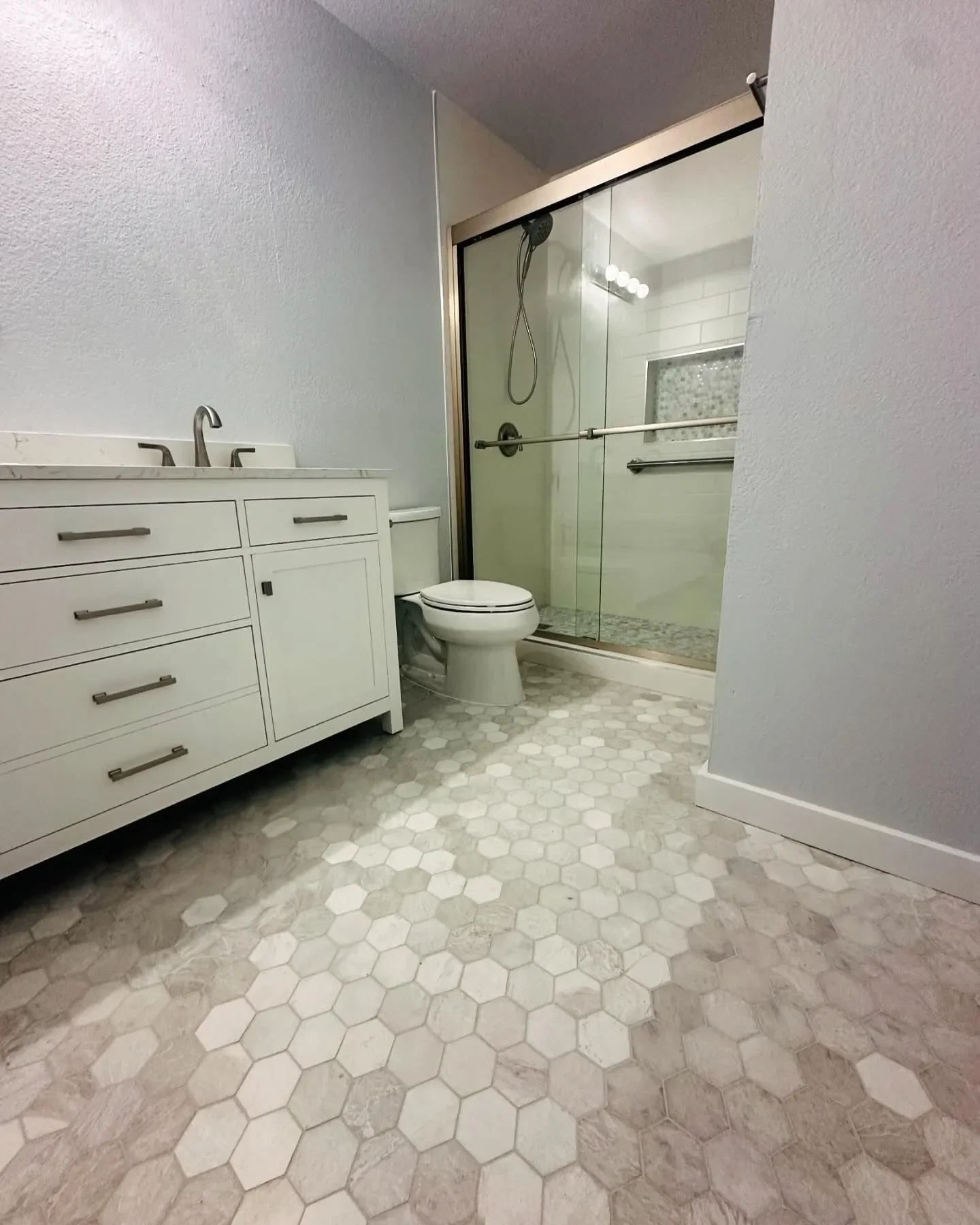A bathroom with a white vanity, a toilet, and a glass shower enclosure with a sliding door and a grab bar. The bathroom has hexagon tile flooring in shades of beige and white.