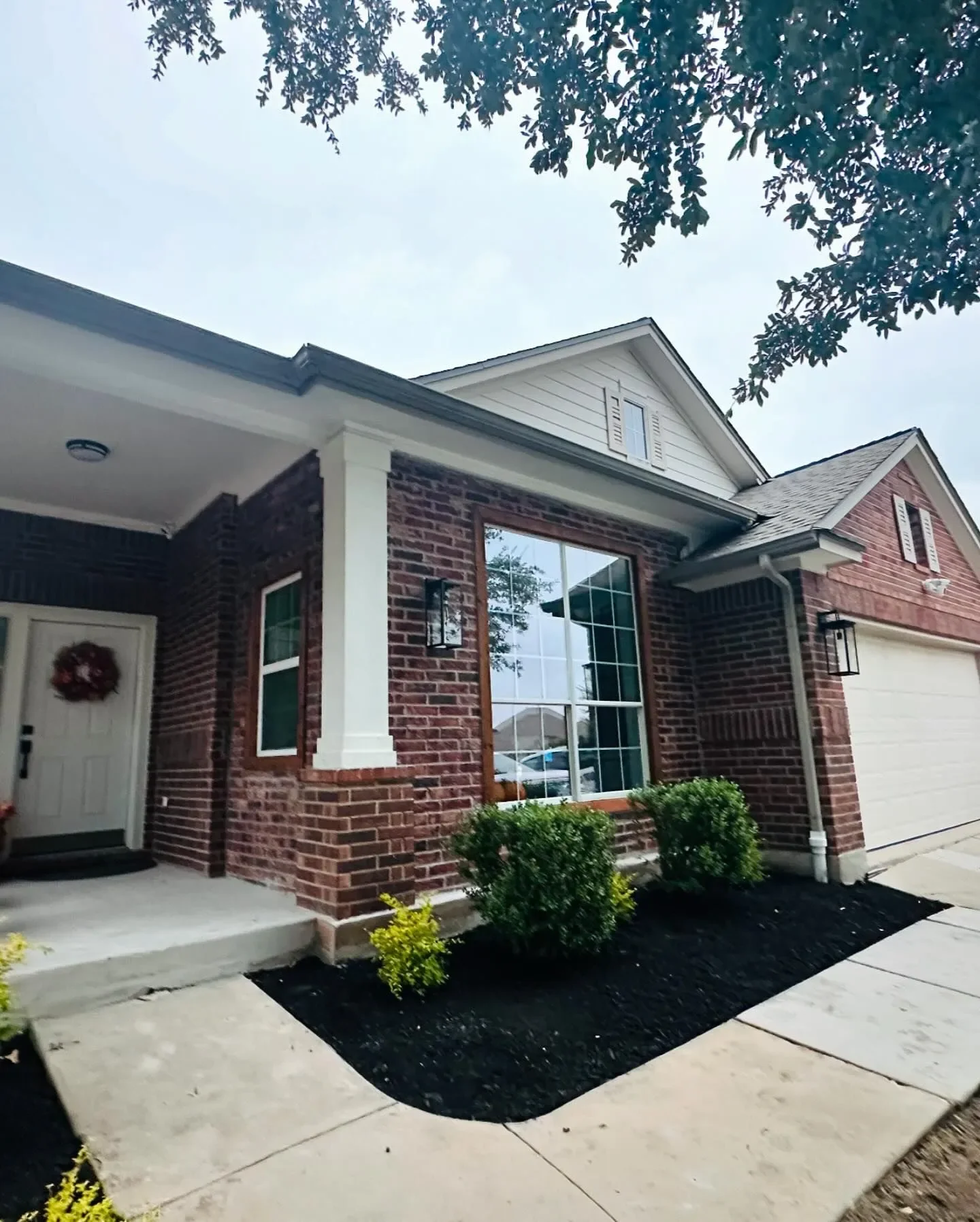 Front view of a brick house with a white garage door, large window, decorated front door with a wreath, landscaped bushes, and a concrete walkway.