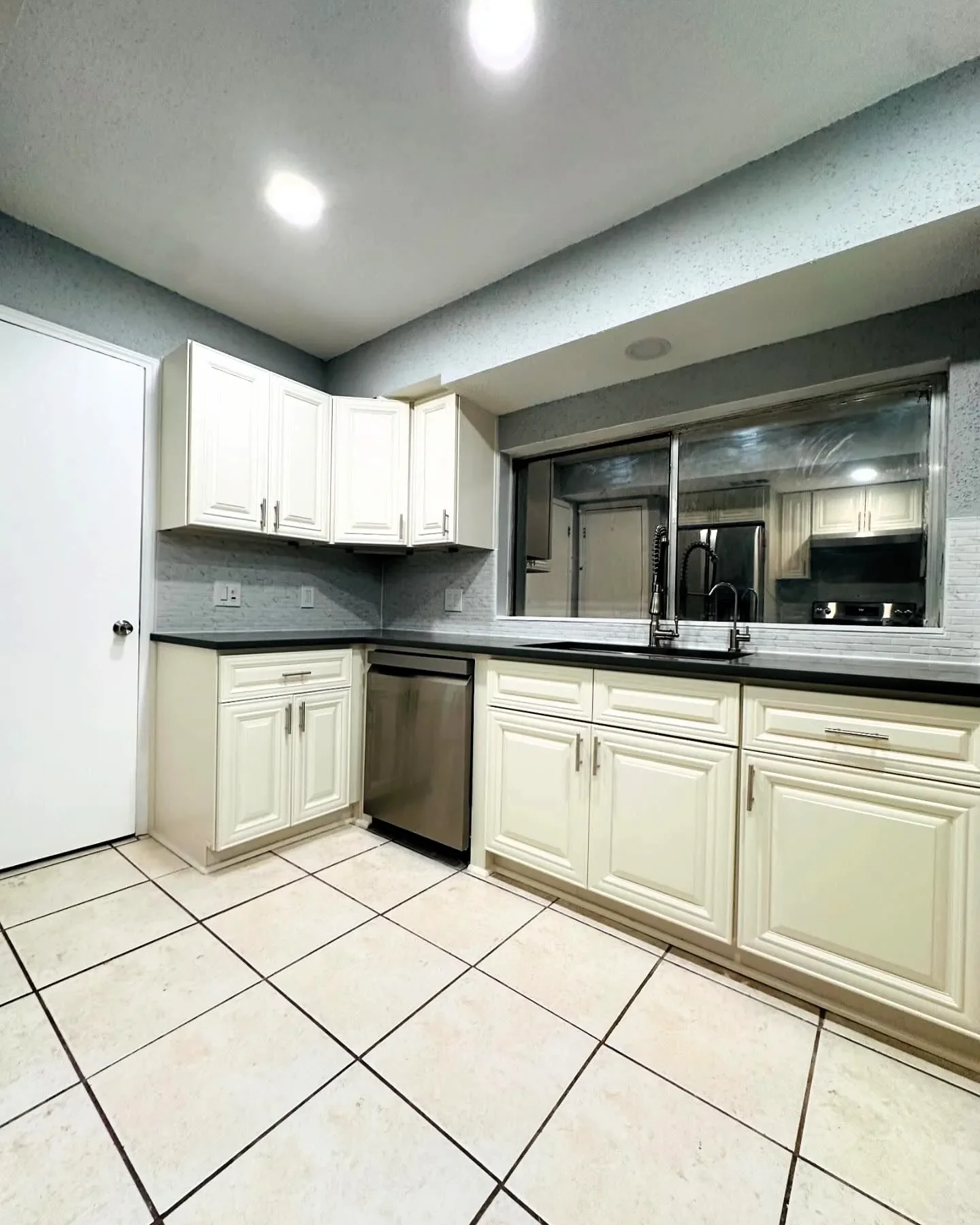 Kitchen with white cabinets, a black countertop, a stainless steel dishwasher, and tiled floor. A large window looks into an additional space with similar cabinetry.