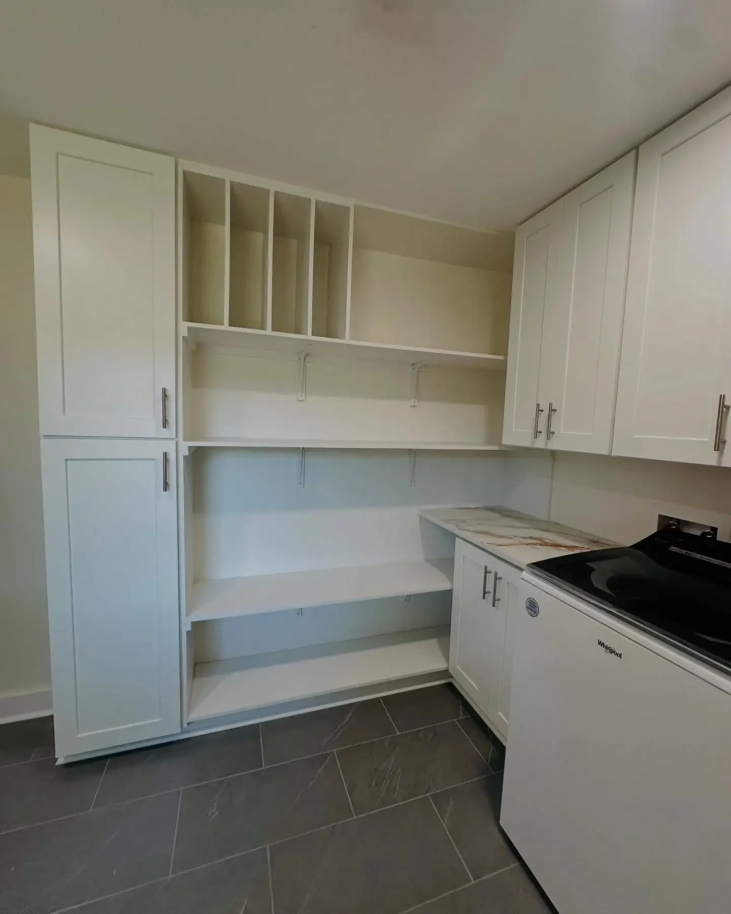 Empty white kitchen cabinet with open shelves, closed cabinets, and a Whirlpool washing machine next to a marble countertop, with dark gray floor tiles.