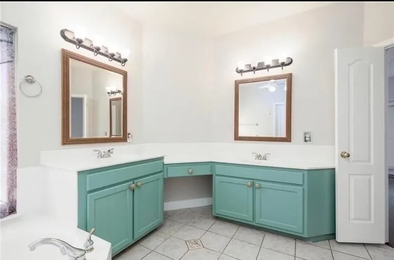 Bathroom with double vanity sinks, two mirrors, and light fixtures, featuring teal cabinets and beige tiled floor.