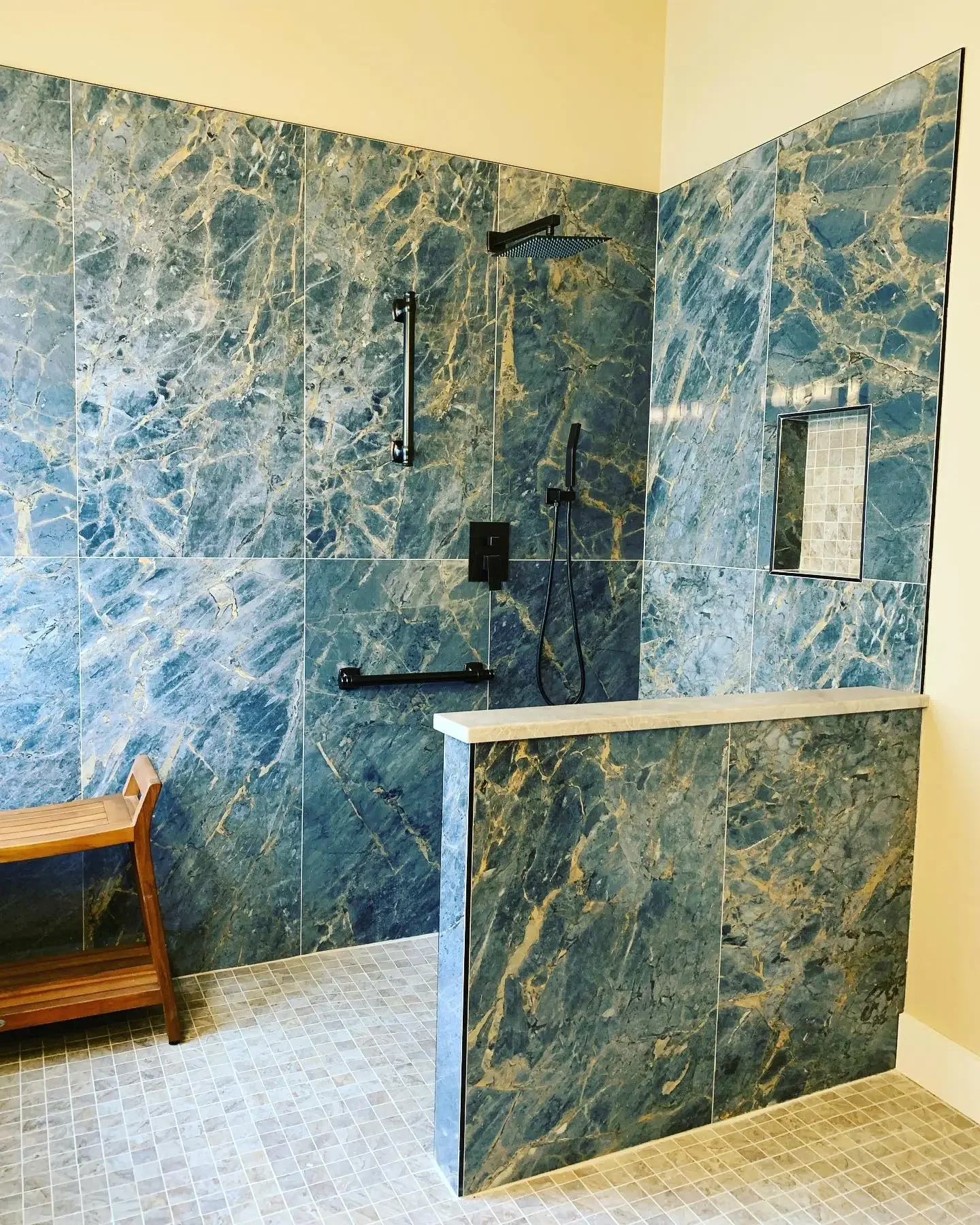 A modern walk-in shower with blue and gold marble tiles, a black rain shower head, a handheld shower, a small inbuilt shelf, a black grab bar, and a partial glass divider.