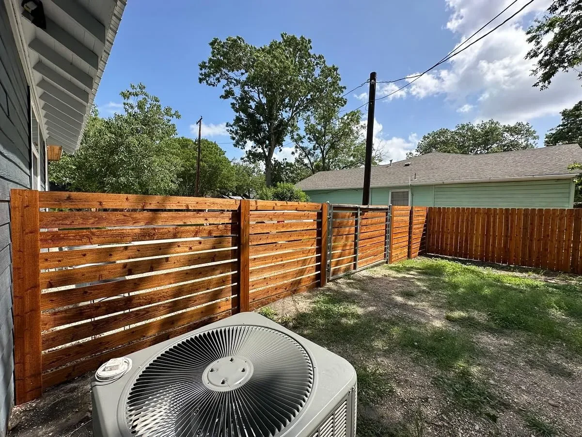 Backyard with a newly installed wooden fence, a yard with grass and dirt, and a large tree in the background. There is an air conditioning unit in the foreground and power lines overhead.