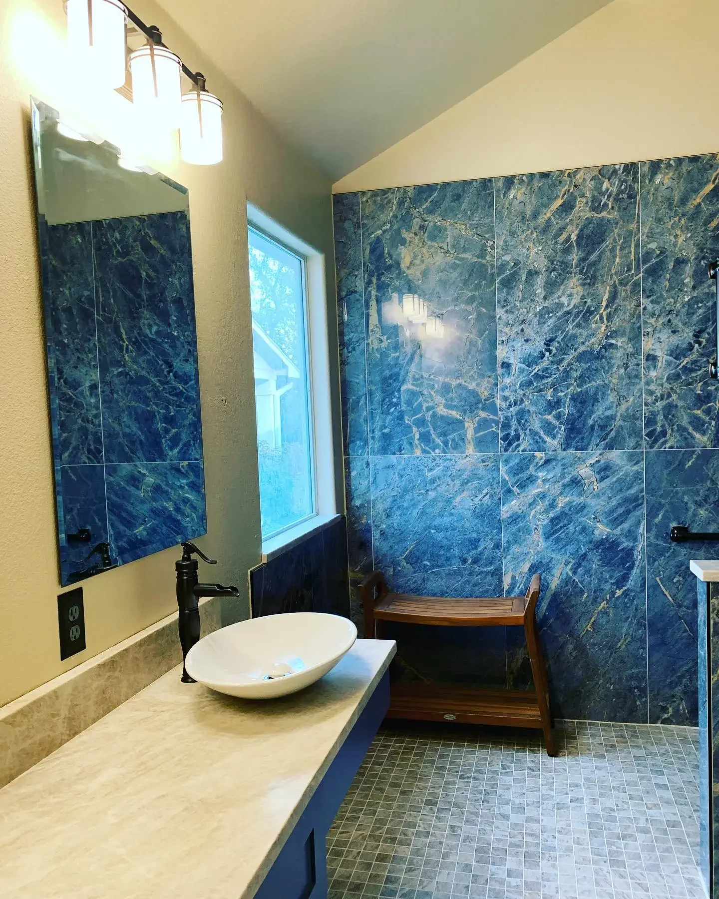 Bathroom with beige countertop, vessel sink, black faucet, large mirror, wood bench, blue marble tile wall, window, and mosaic tile floor.