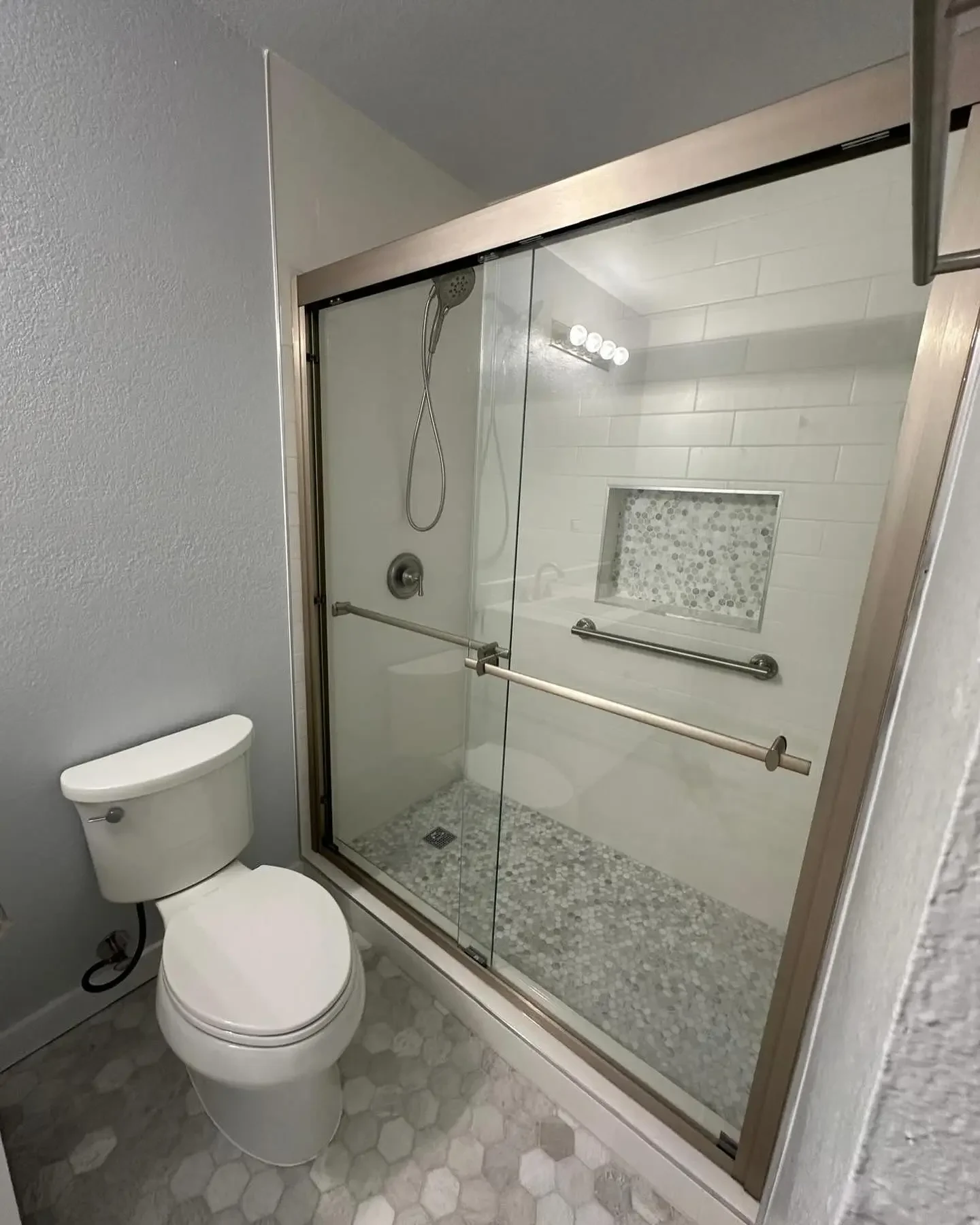 A bathroom with a toilet and a glass-enclosed shower featuring a built-in niche, grab bars, and a pebble stone shower floor.