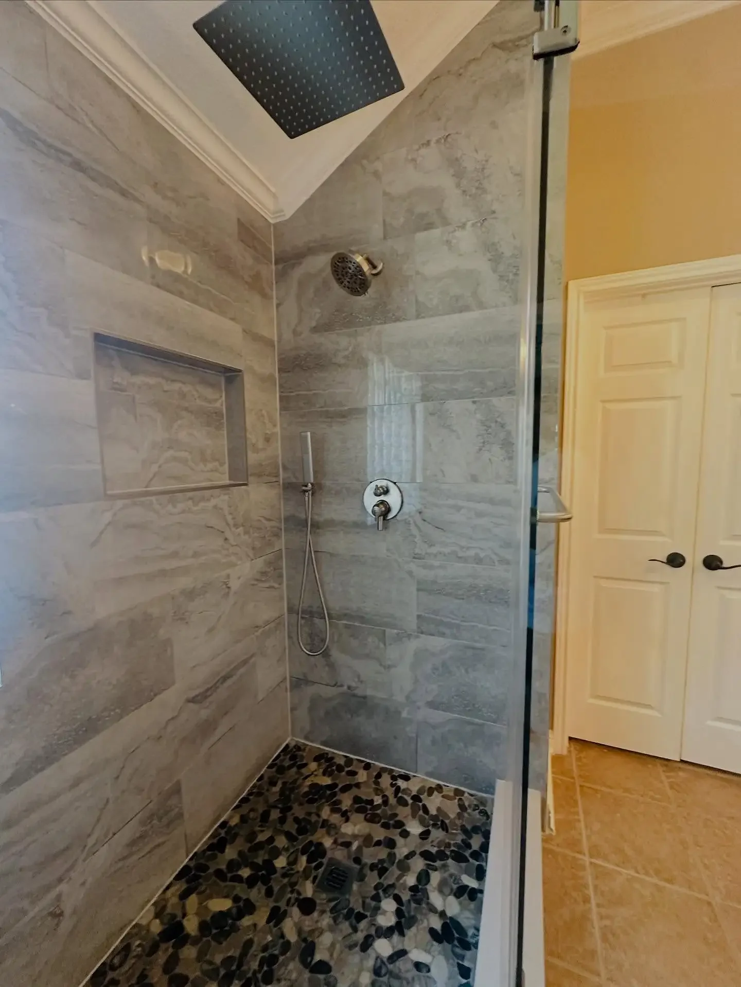 Shower with gray marble tiles, a black pebble tile floor, a ceiling-mounted black square rain shower head, a wall-mounted adjustable hand shower, and a built-in niche for toiletries.