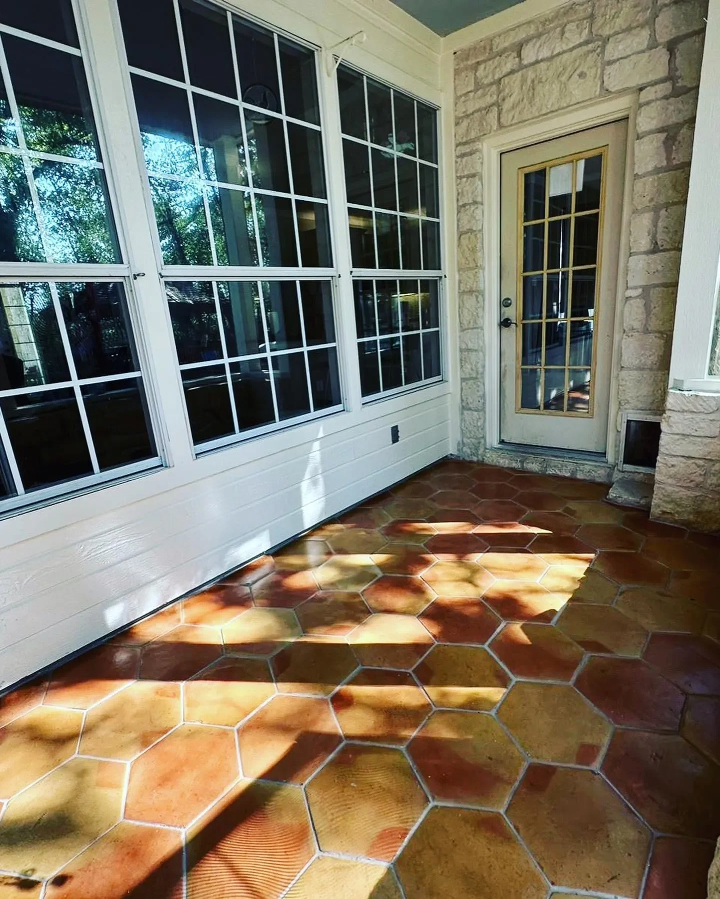 A porch area with large windows, a door with windowpanes, and a terracotta tile floor with sunlight and shadow patterns.