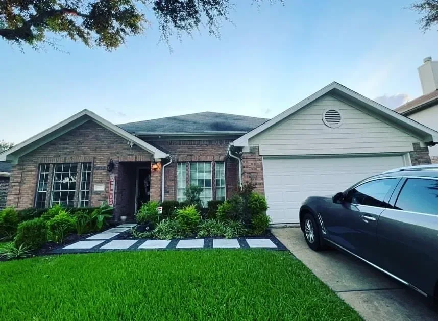 A single-story house with a brick and white siding exterior, a manicured front lawn with lush green grass, a pathway leading to the front door, and a black car parked in the driveway under a clear blue sky.