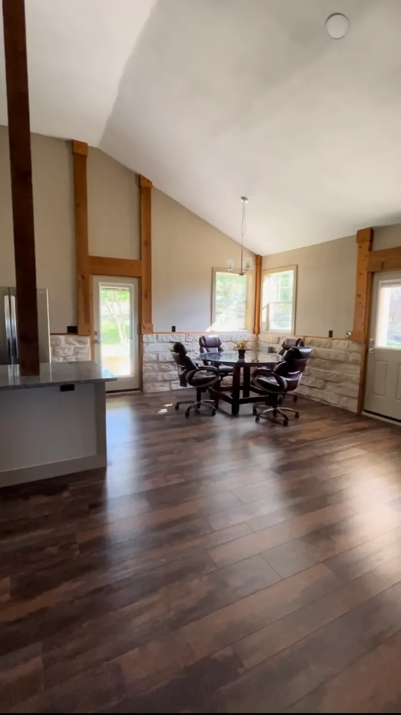 Open concept dining area with dark wood flooring, four black office chairs around a round table, beige stone half-wall, and large windows with views of greenery, wooden beams, and a sloped ceiling.