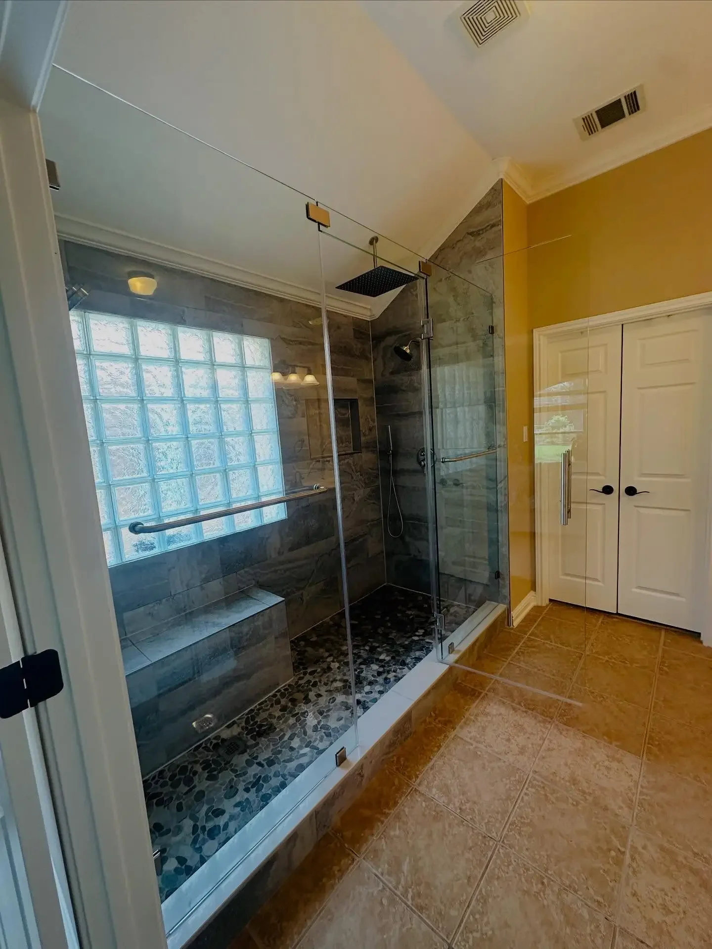 A modern walk-in shower with glass enclosure, dark stone tile walls, pebble tile flooring, and a built-in bench. There is a large square rain showerhead and a smaller handheld shower, with a pull-up bar on one wall. The bathroom has yellow walls, a t