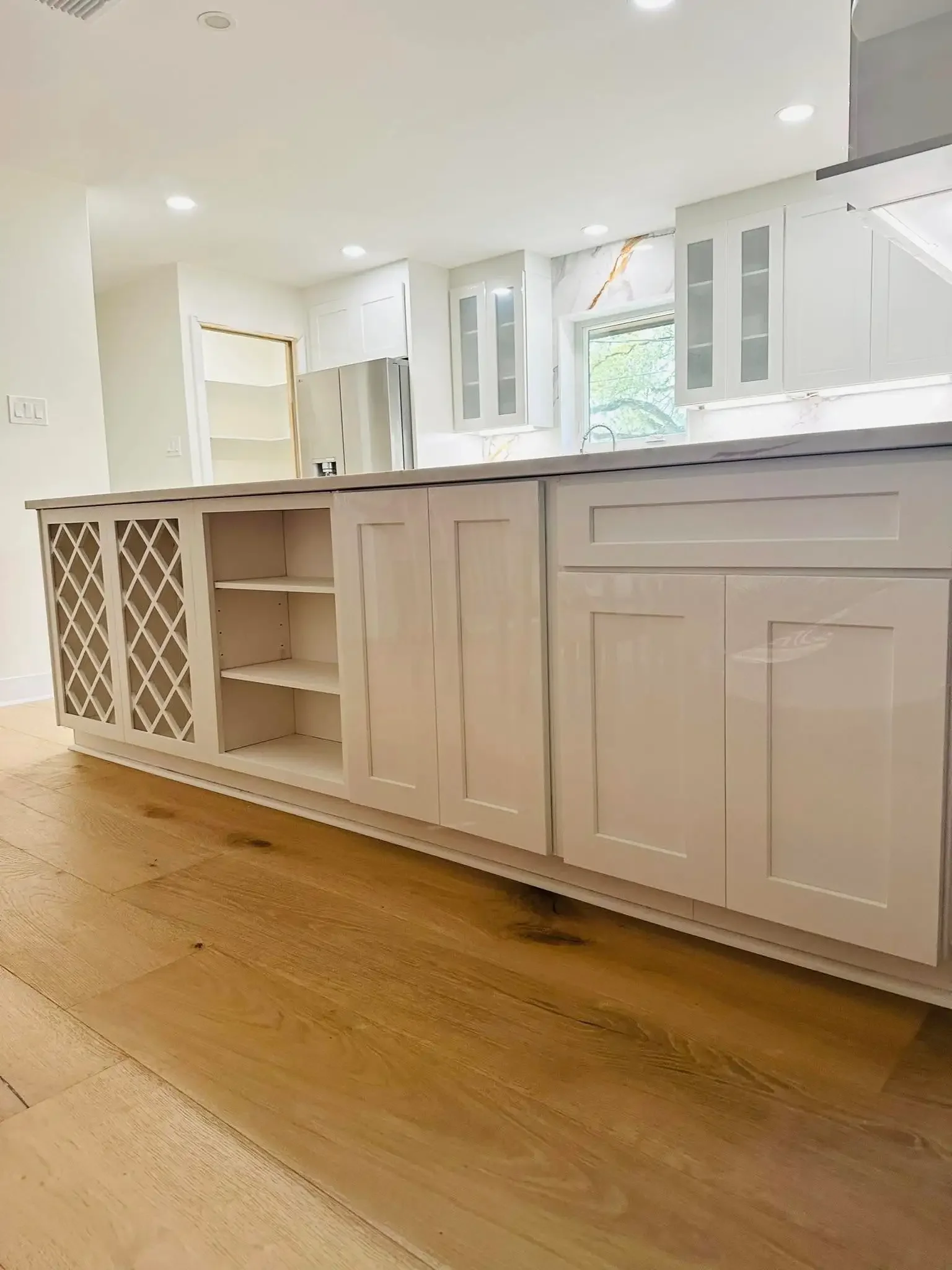 Modern kitchen with white cabinets, a marble backsplash, and a wooden floor. The kitchen island has open shelving and a wine rack, with a window above the sink.
