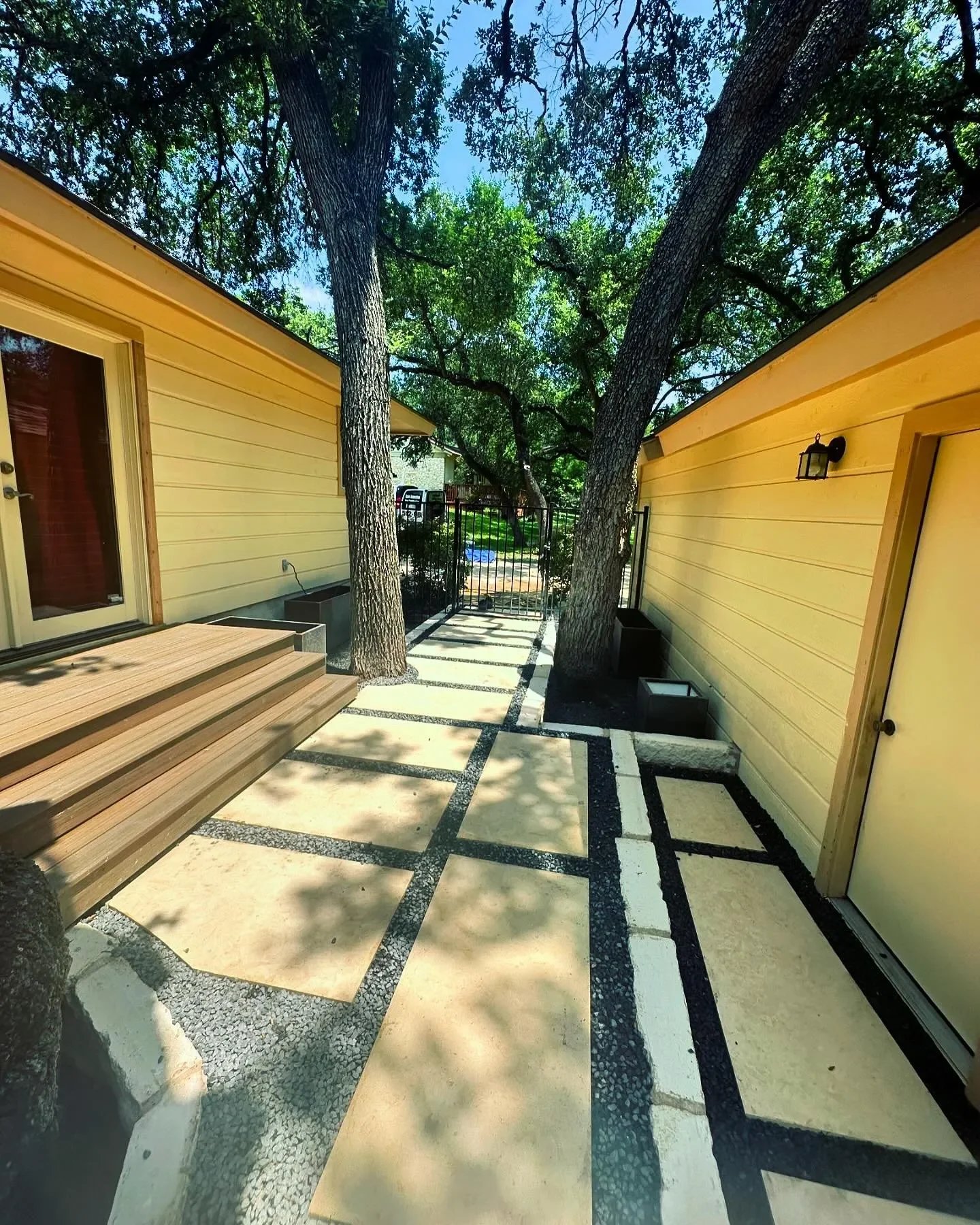 A backyard patio with large trees providing shade, yellow house siding on both sides, a sliding glass door with wooden steps on the left, a black metal gate at the end of the patio, and a paved pathway with a geometric black and white pattern.